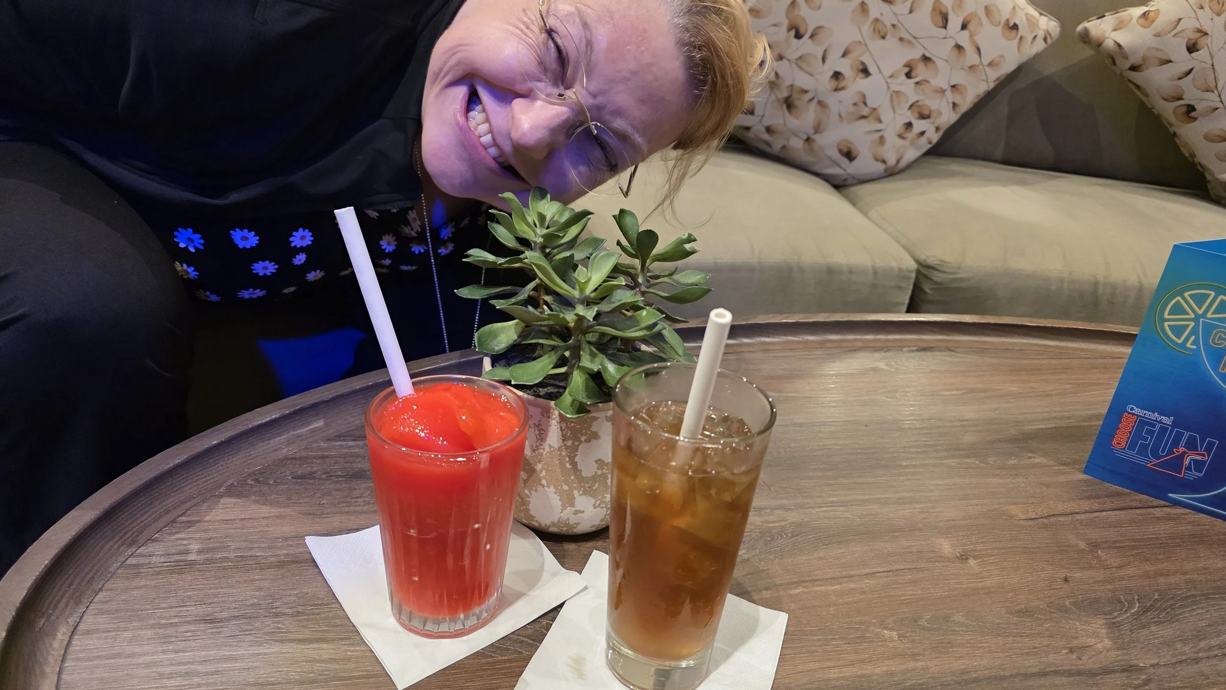 A woman with glasses smiling and leaning over a wooden table with two drinks, a planter with a green succulent, and a blue card on it in a cozy indoor setting.