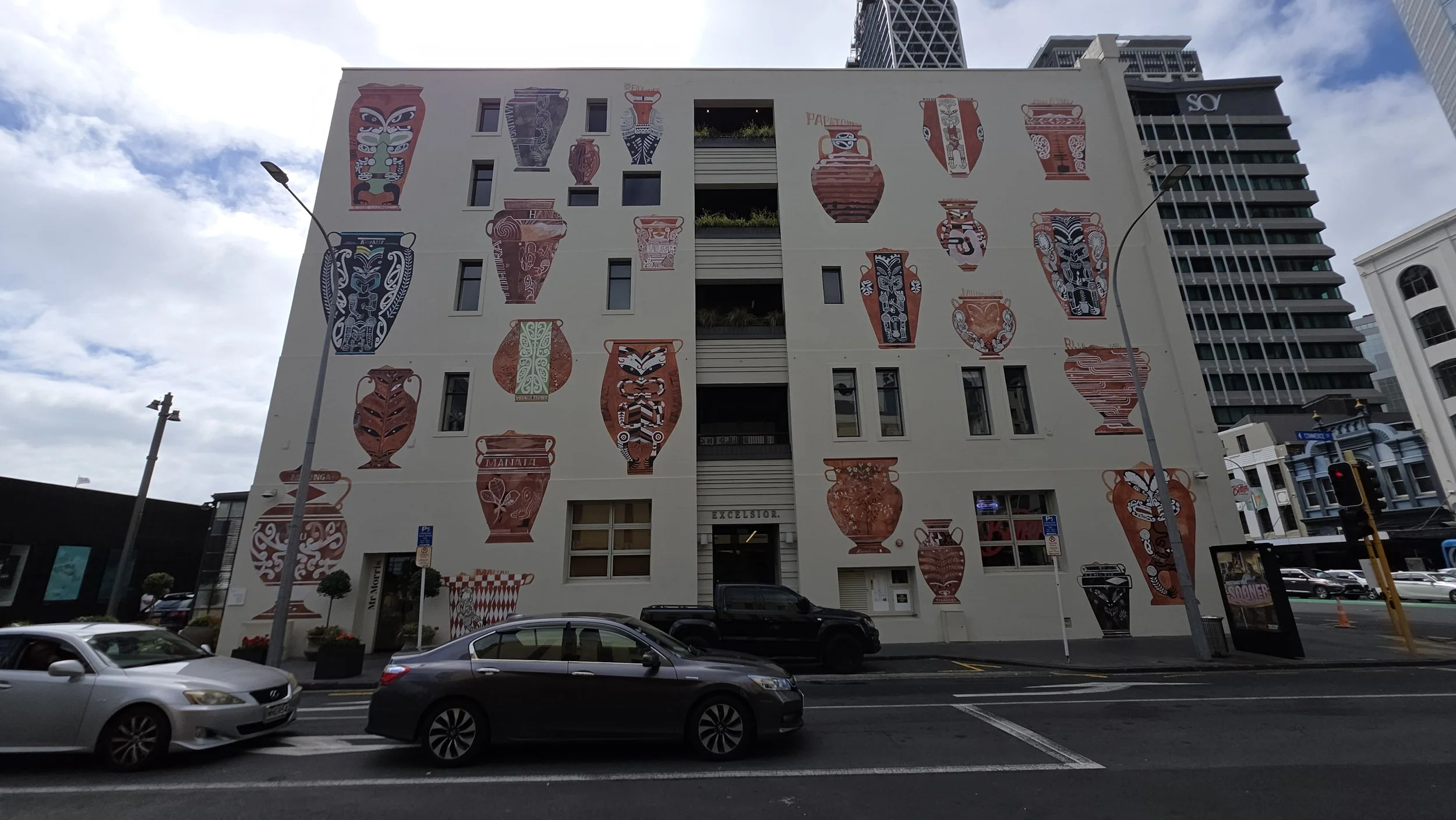 A large white building with murals of various colorful, decorative pottery and vases painted on its facade, situated on a city street with cars and street lamps.