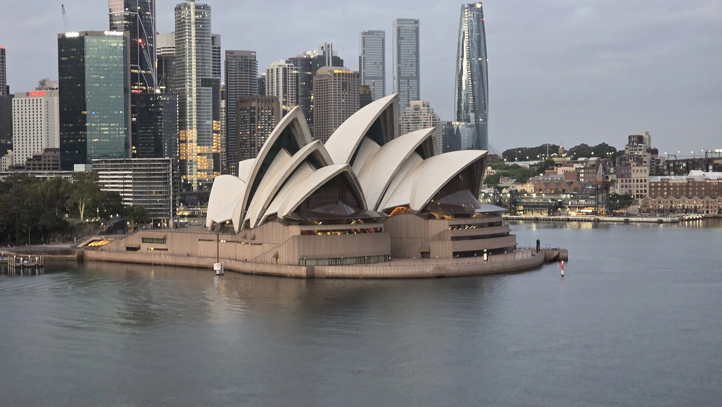 Sydney Opera House with city skyline in the background at dusk, reflecting on calm water.