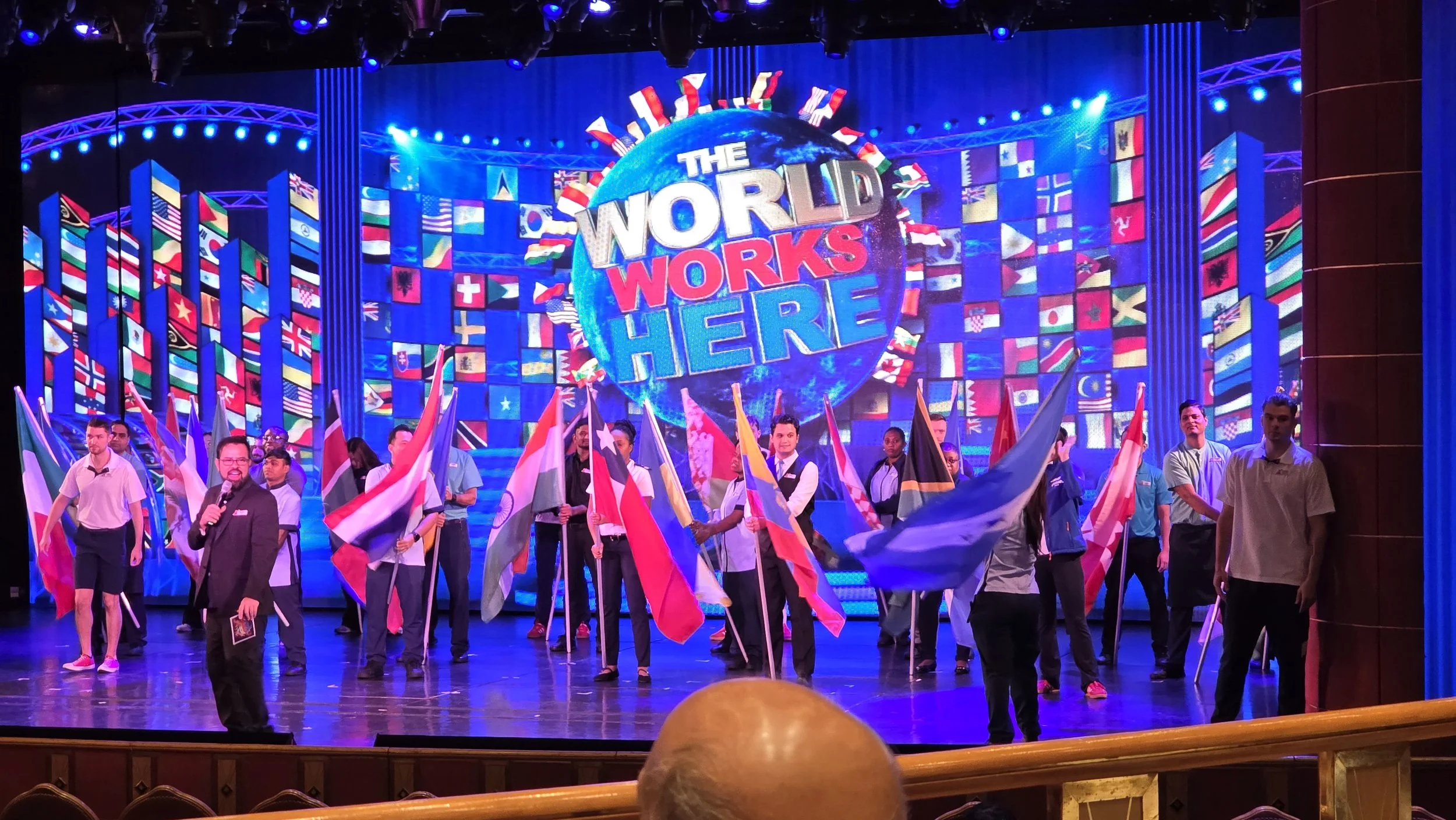 Group of people on stage holding various international flags, in front of a large screen displaying the text 'The World Works Here' with many international flags in the background.