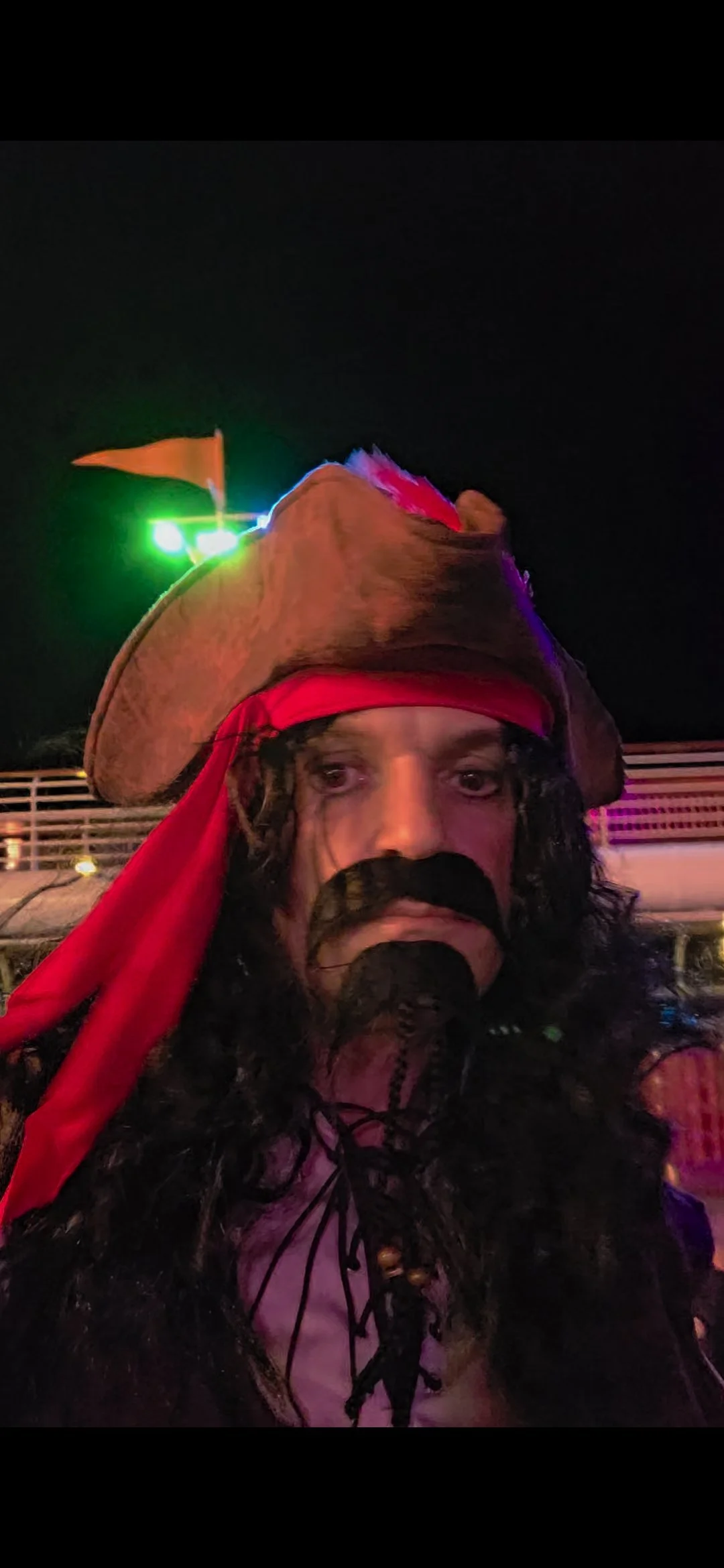 Person dressed as a pirate, wearing a brown hat with a red band and a small orange flag, with long curly dark hair, a black mustache and beard, holding a microphone, at night with colorful lights in the background.