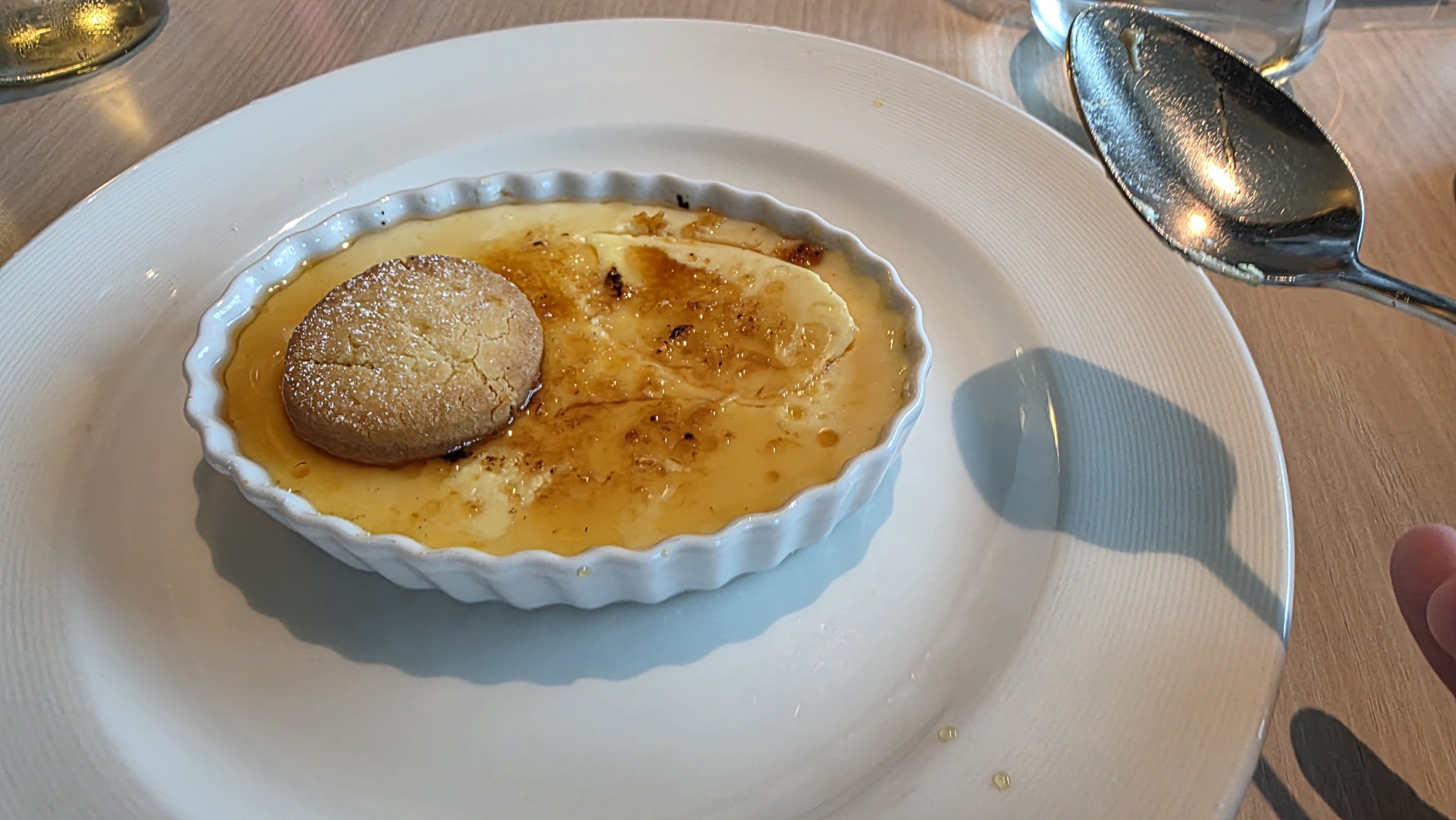A ramekin of creme brulee topped with a cookie on a white plate, with a spoon and a glass of water nearby.