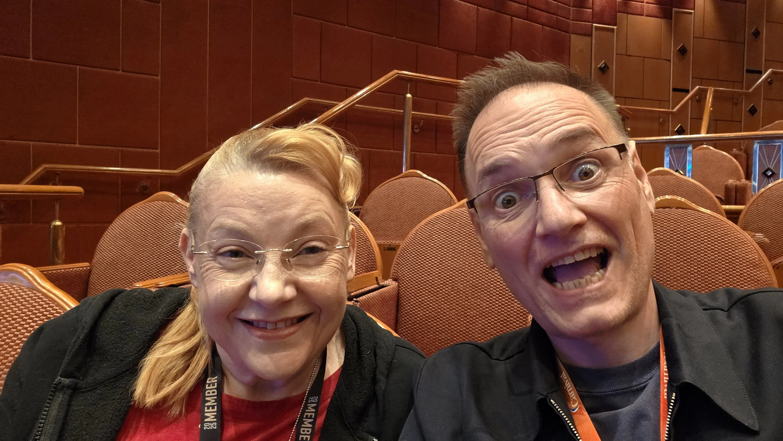A woman with glasses and red hair next to a man with glasses in a theater or auditorium with red velvet seats. They are smiling and looking at the camera.