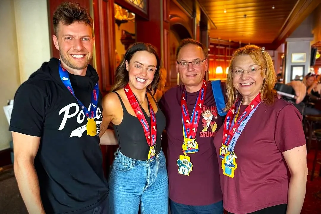 Four people smiling and wearing medals at an indoor event, with wooden walls and other people in the background.
