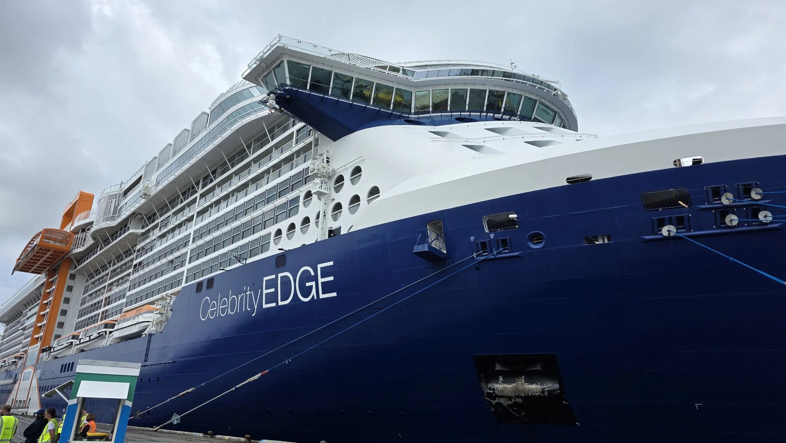 A large cruise ship named Celebrity EDGE docked at a port, with people in safety vests on the dock and overcast sky in the background.