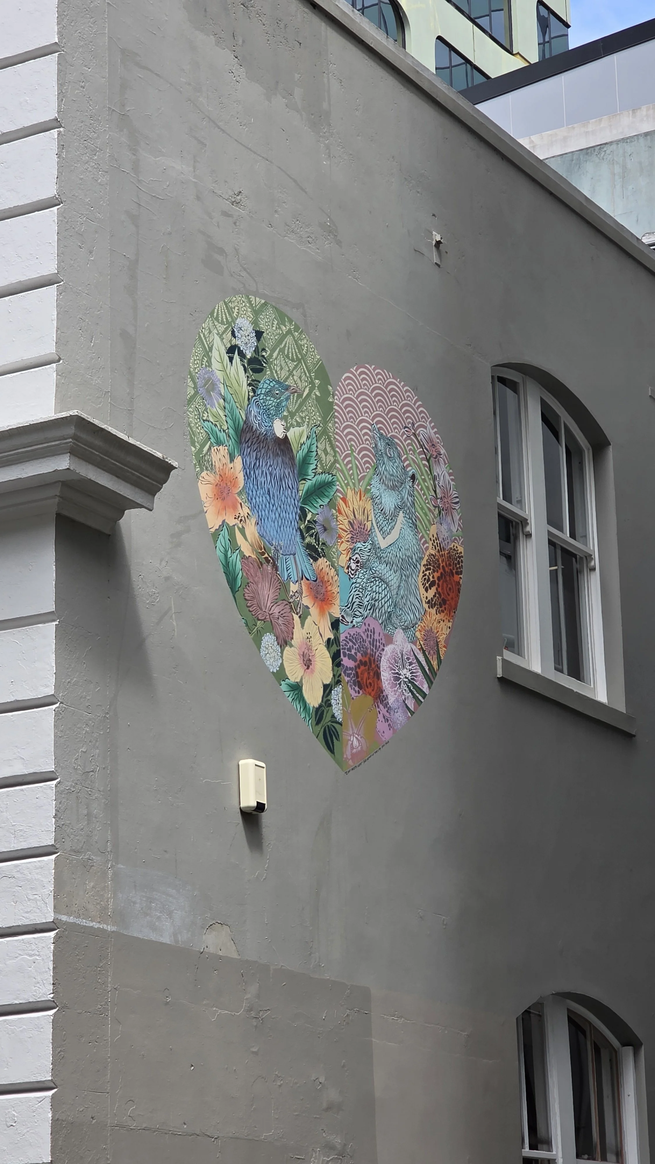 A mural of colorful flowers and two birds inside a heart shape on the side of a building.
