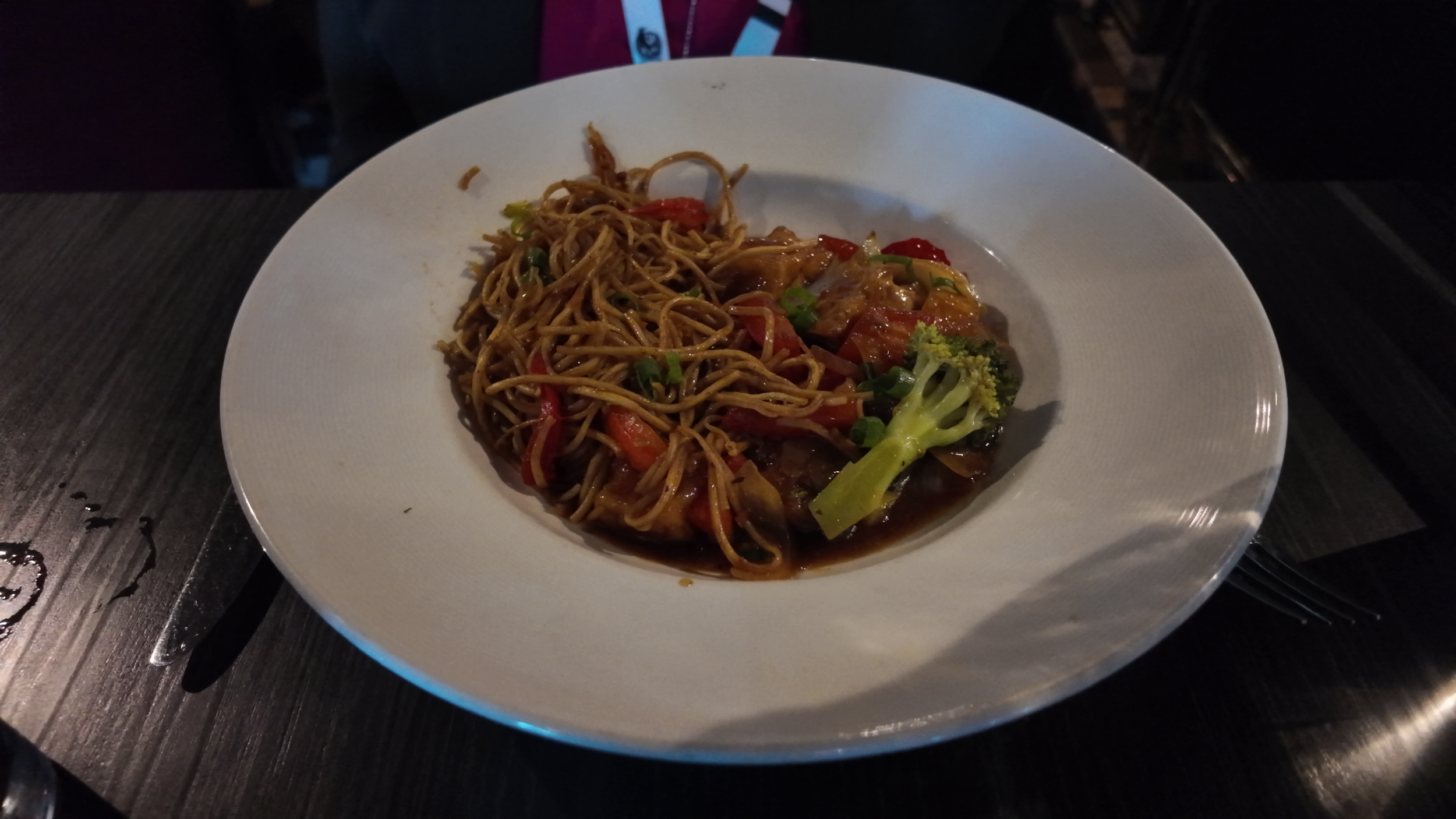 A white plate with Asian-style noodles and vegetables, including red peppers and broccoli, served with soy sauce on a dark wooden table.