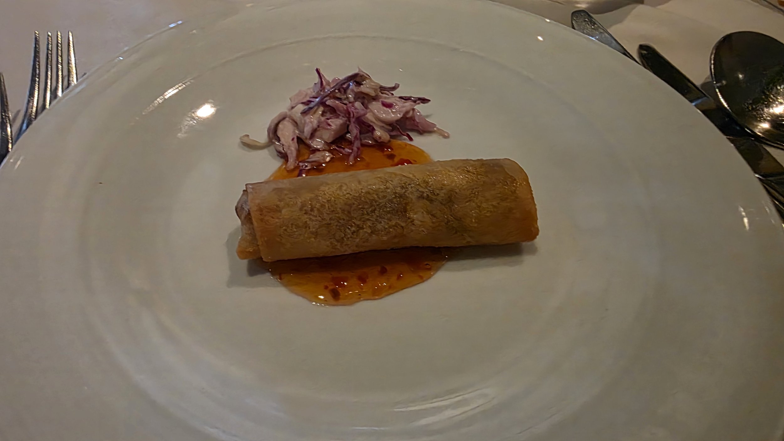 A plated appetizer with a spring roll on a large white plate, accompanied by a small portion of shredded meat and a sauce.