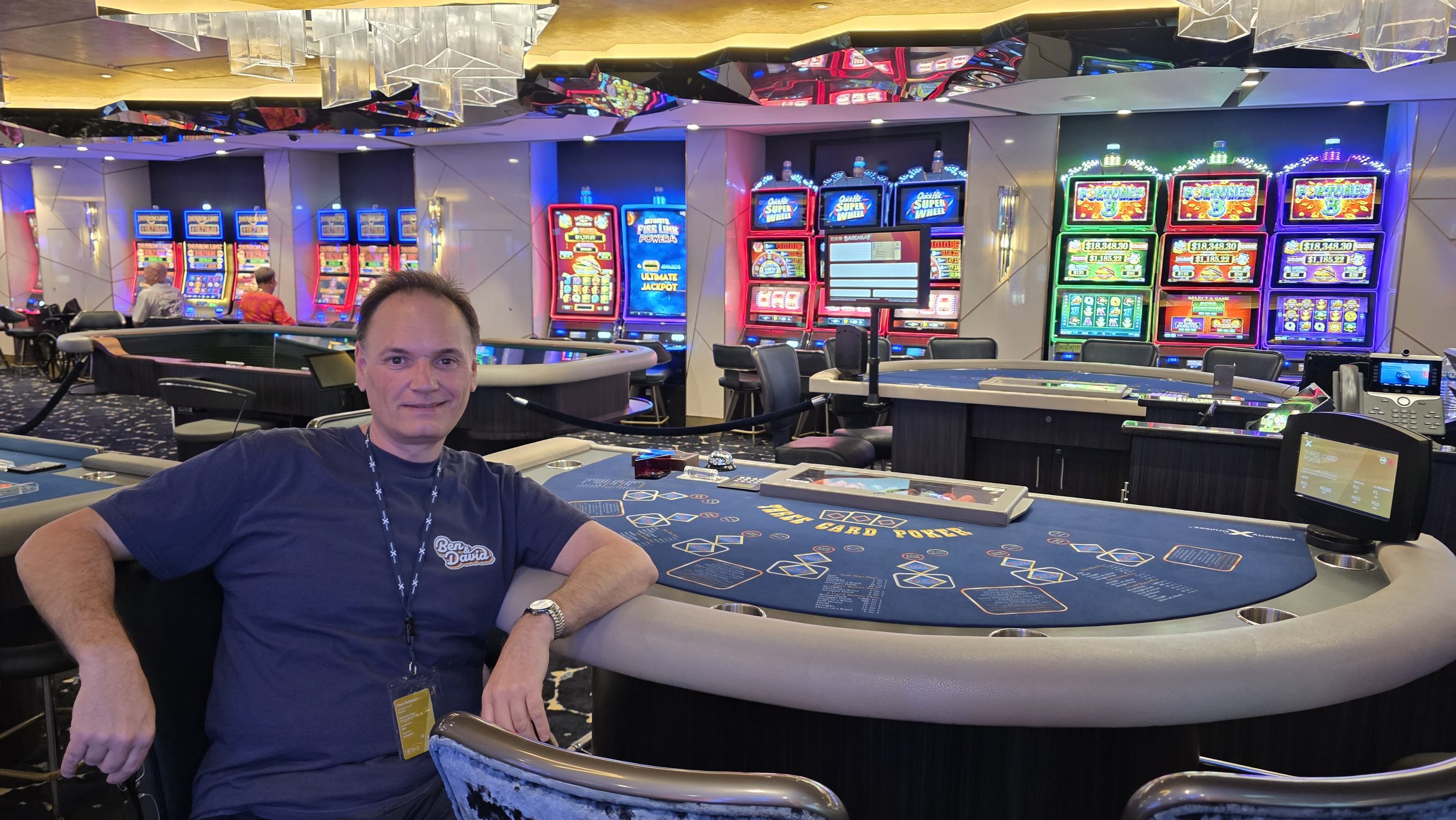 A man sitting at a poker table inside a casino with colorful slot machines in the background.