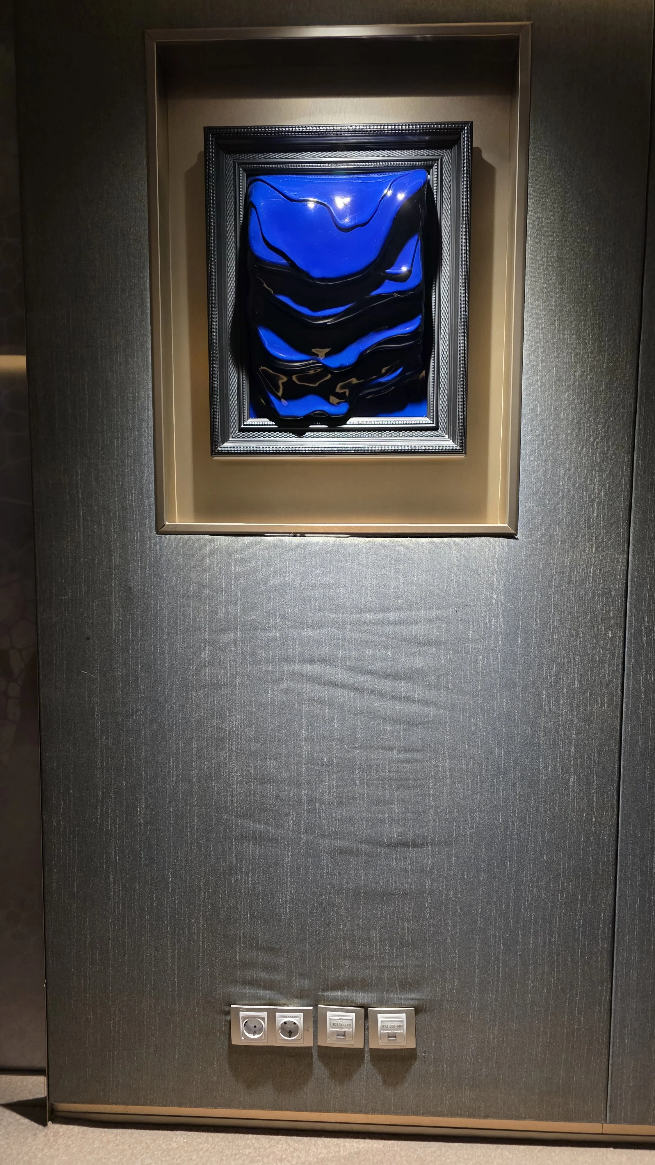 An abstract artwork featuring swirling blue and black patterns, framed and hung on a dark textured wall with two power outlets and switches beneath.