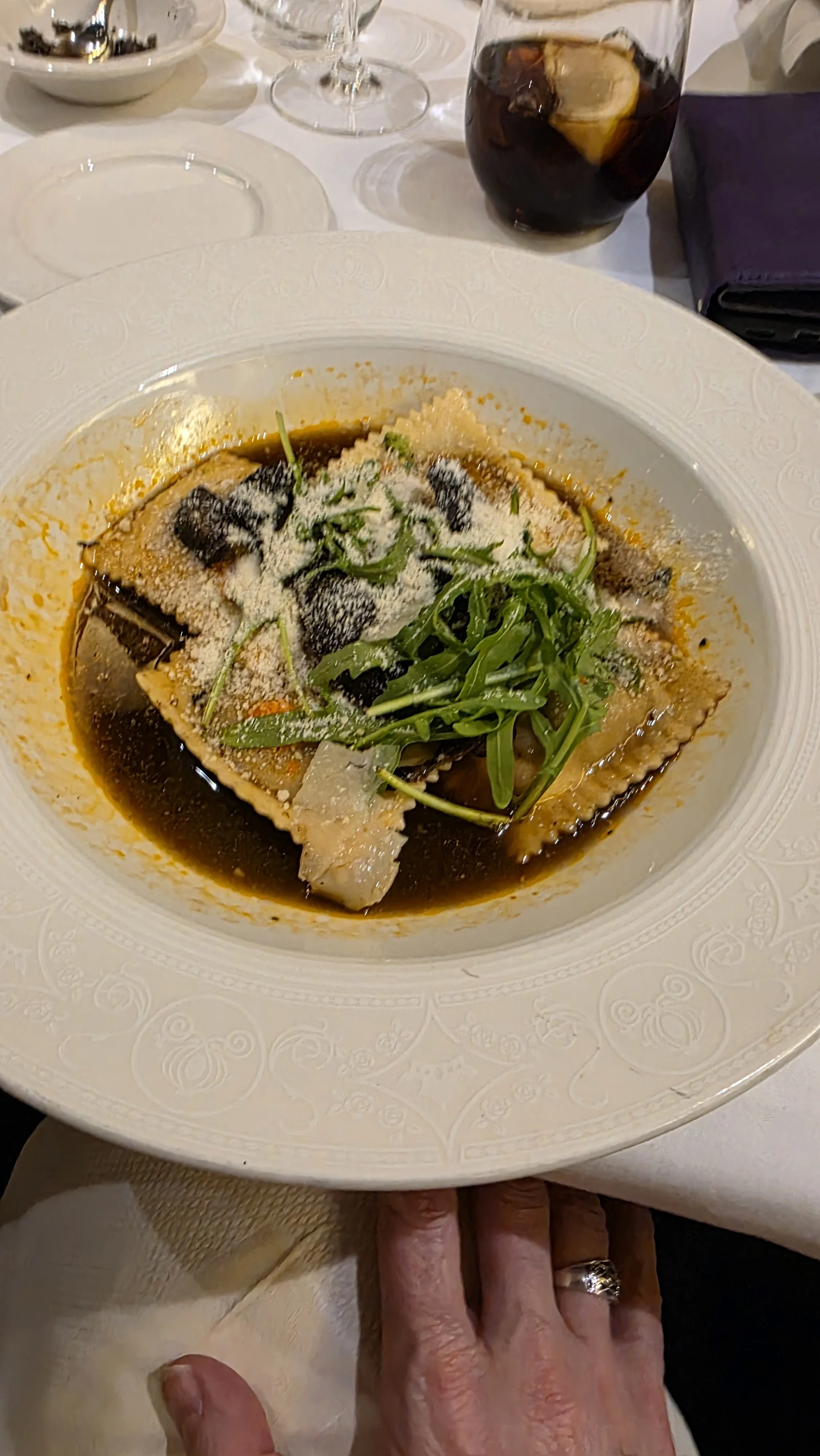 Plate of tortellini pasta topped with grated cheese and arugula, served in a dark broth or sauce. In the background, there is a glass of soda with lemon and some wine glasses.