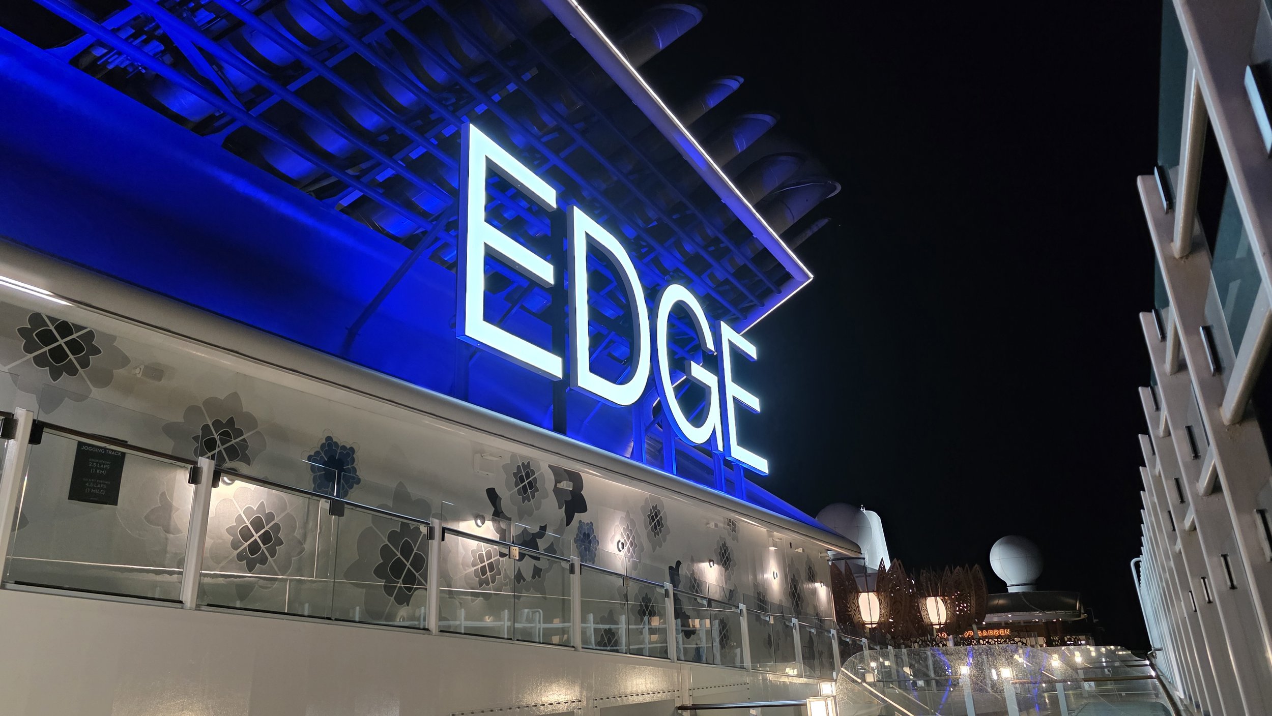 Nighttime view of the side of a cruise ship with a large illuminated sign that reads 'EDGE', blue and white lighting, windows, decorative wall panels with flower-like patterns, and other structures on the ship's deck.