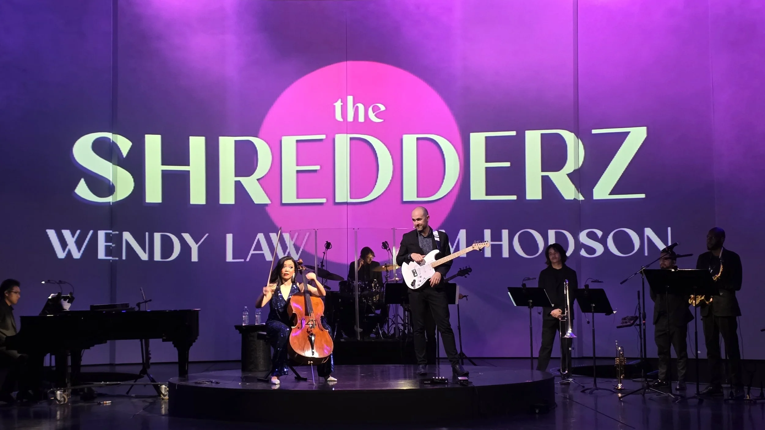 A musical performance on stage with a large screen in the background displaying the text: 'the SHREDDERZ WENDY LAW RUTH MORPDSON.' There is a woman playing a cello, a man playing an electric guitar, and other musicians with instruments.