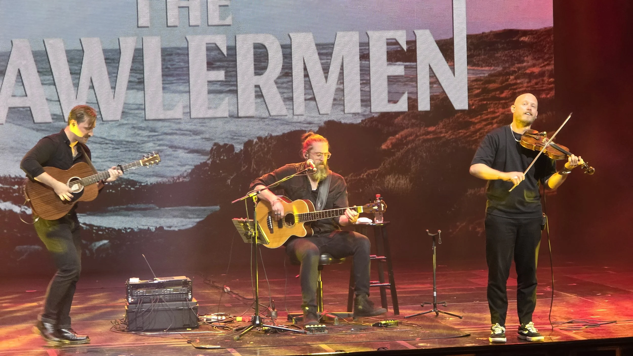 Three musicians performing on stage with guitars, with a large screen behind them displaying the words 'THE AWLERMEN' and a background image of a landscape.