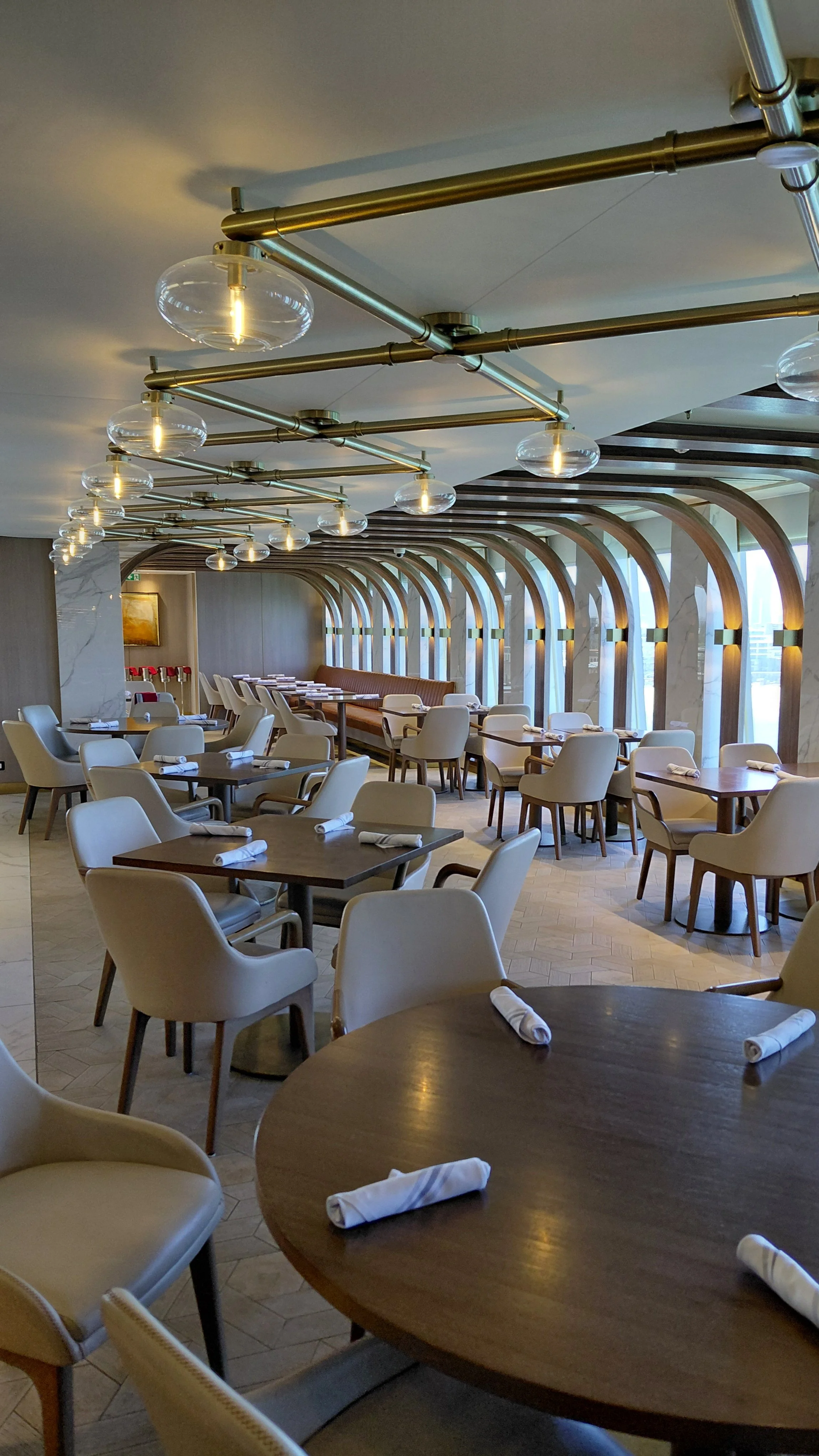 Empty restaurant with wooden tables, white upholstered chairs, and large arched windows, decorated with modern lighting fixtures.