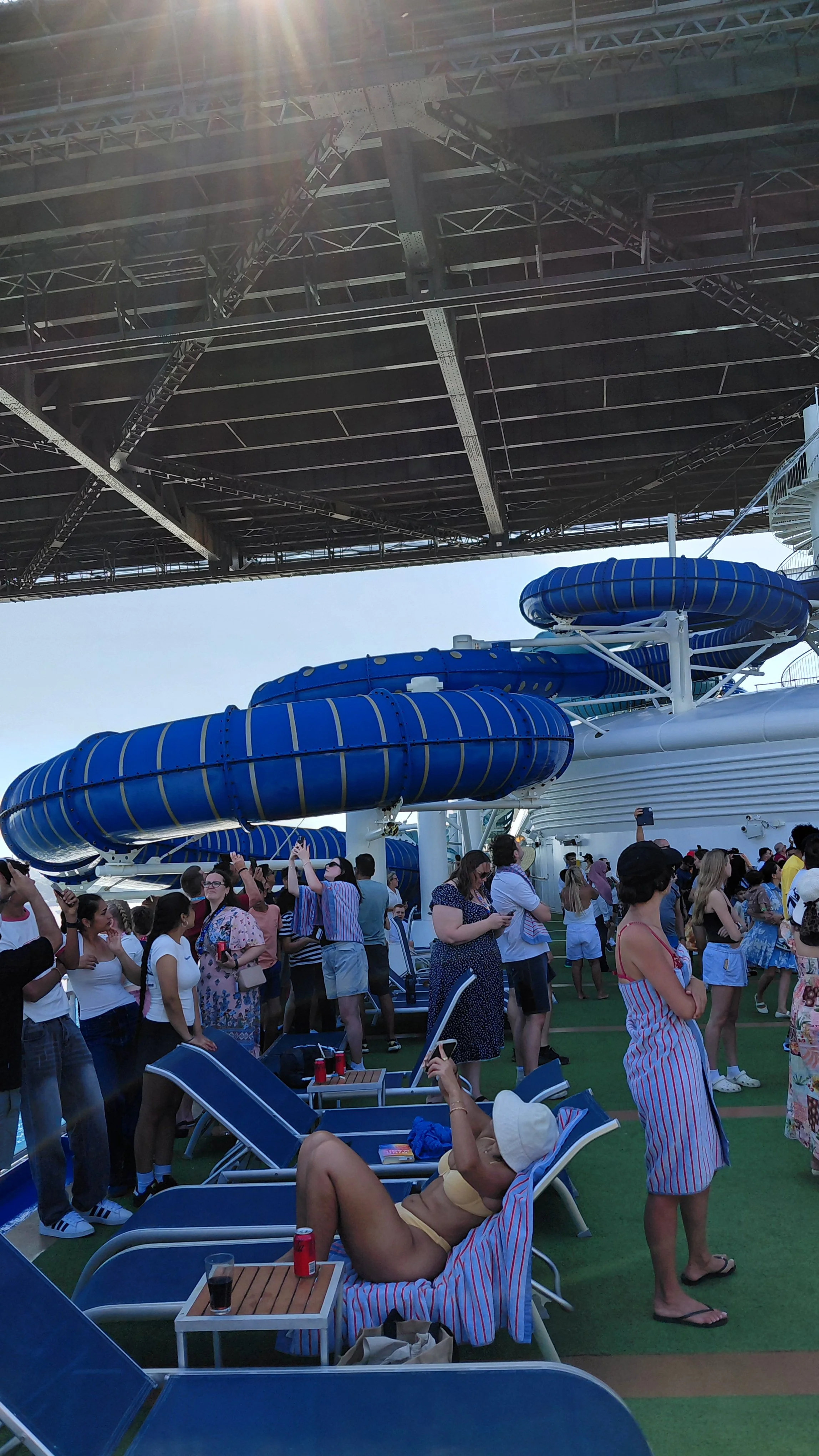 People on the deck of a cruise ship with a large blue water slide and numerous loungers, some people taking photos and relaxing.