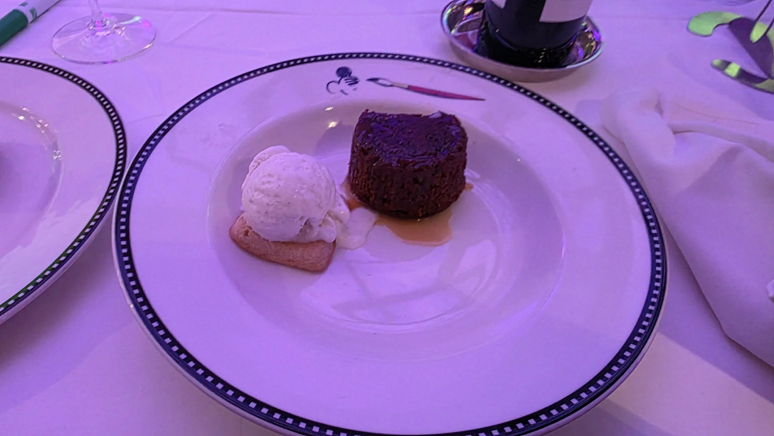 Chocolate lava cake with a side of vanilla ice cream on a white plate with a themed decorative black and white border, set on a white tablecloth, with a glass of wine and a cup in the background.