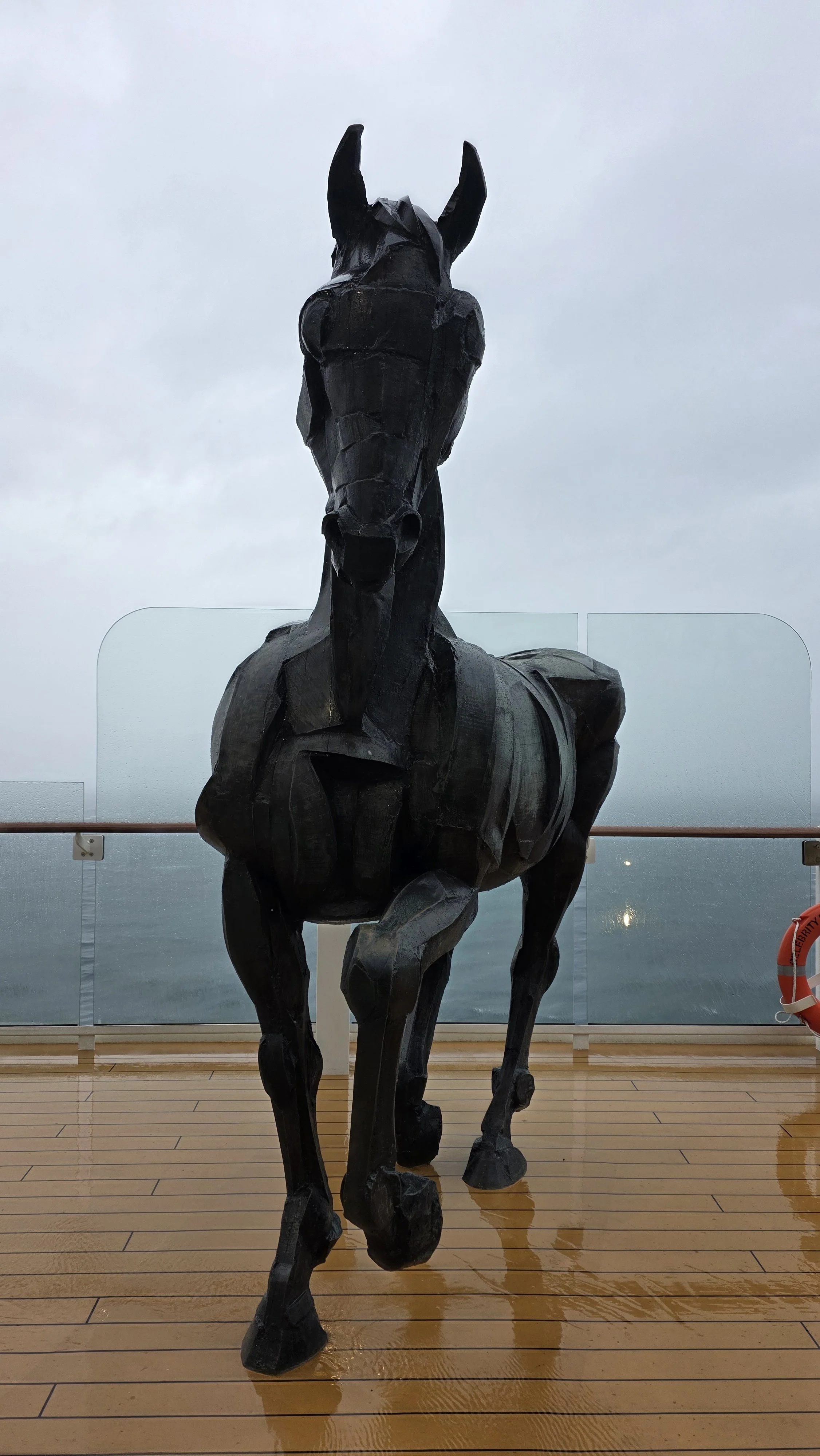 A large black abstract sculpture of a horse on a deck overlooking the ocean.