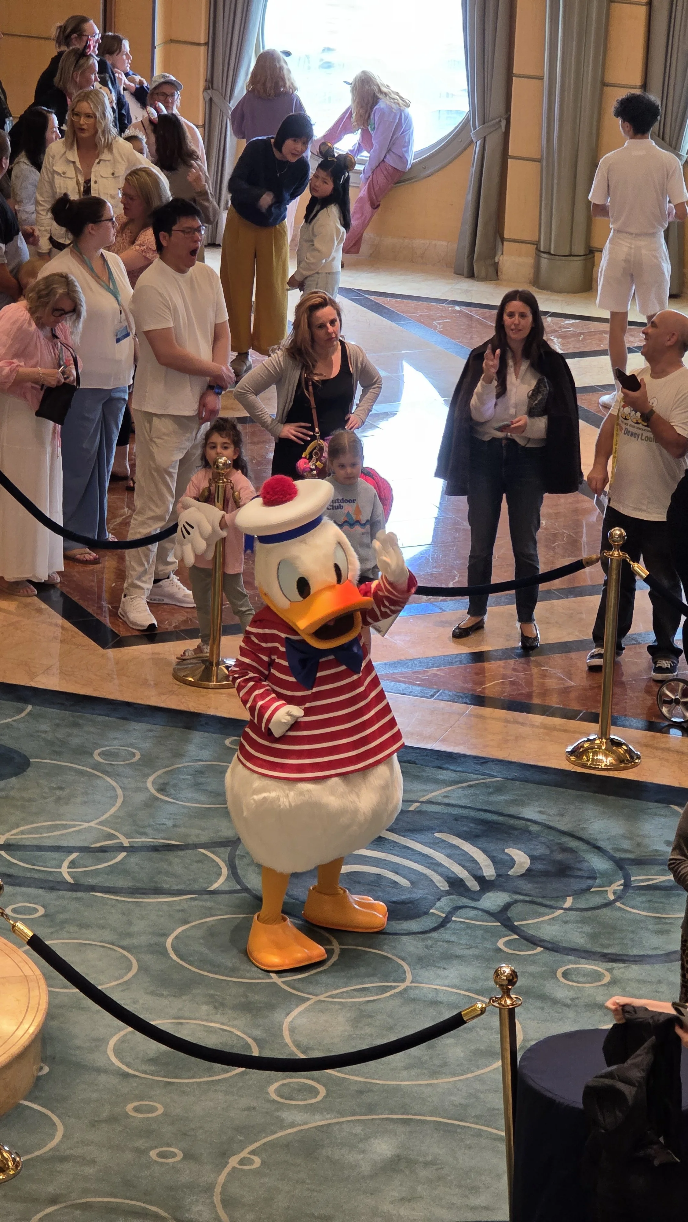 People gather around Disney's Daisy Duck character in a red and white striped shirt and orange shoes, waving at the crowd inside a lobby with marble floors and tall windows.