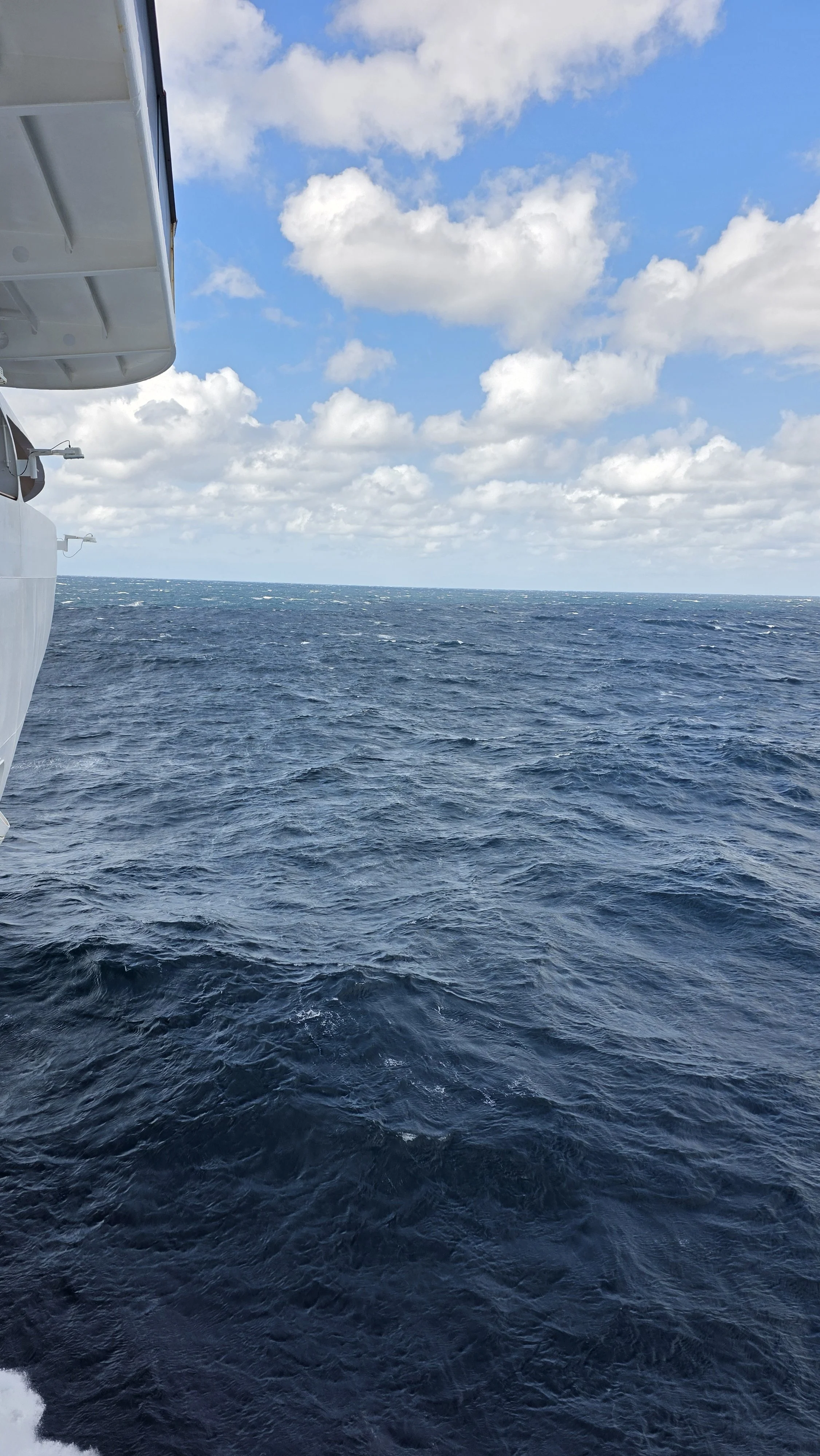 Open ocean view with white clouds in the blue sky, part of a ship's exterior visible on the left.