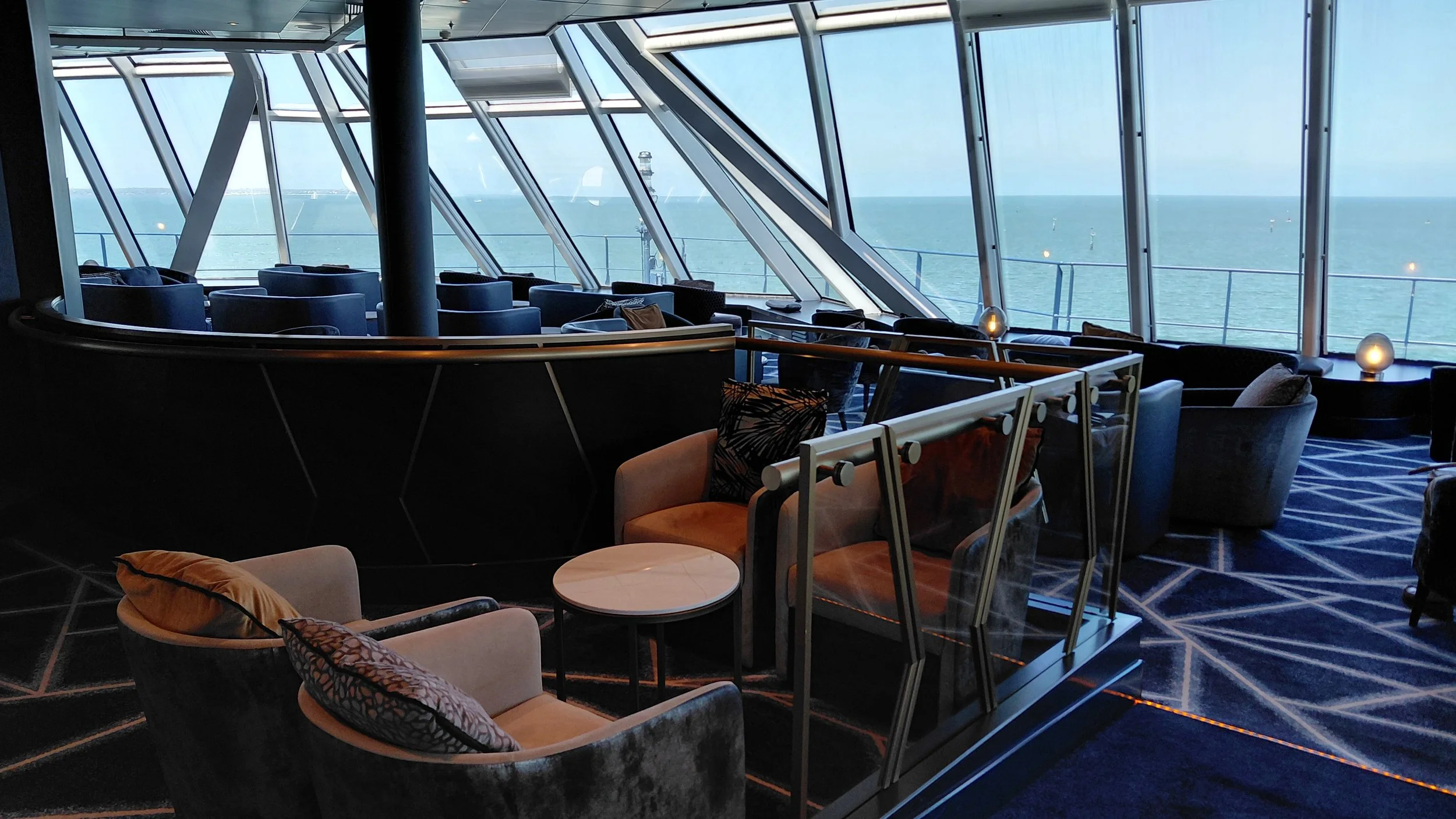 Interior of a lounge area on a cruise ship with large windows overlooking the ocean, modern chairs and tables, soft lighting, and a patterned carpet.