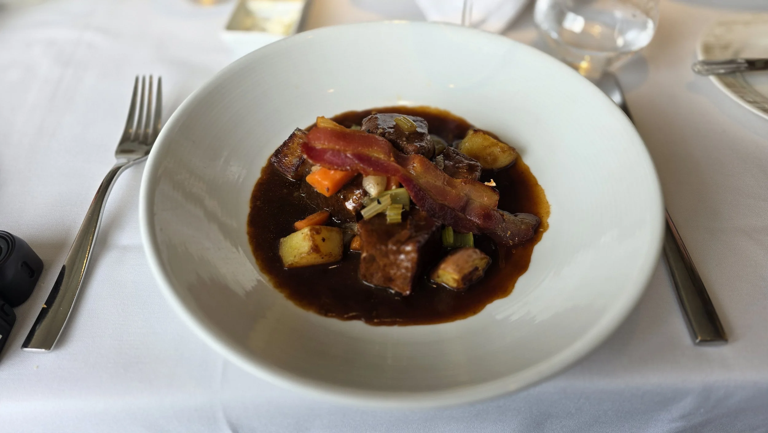 Bowl of beef stew with bacon, potatoes, carrots, celery, and gravy on a white tablecloth, with fork, knife, and glass nearby.