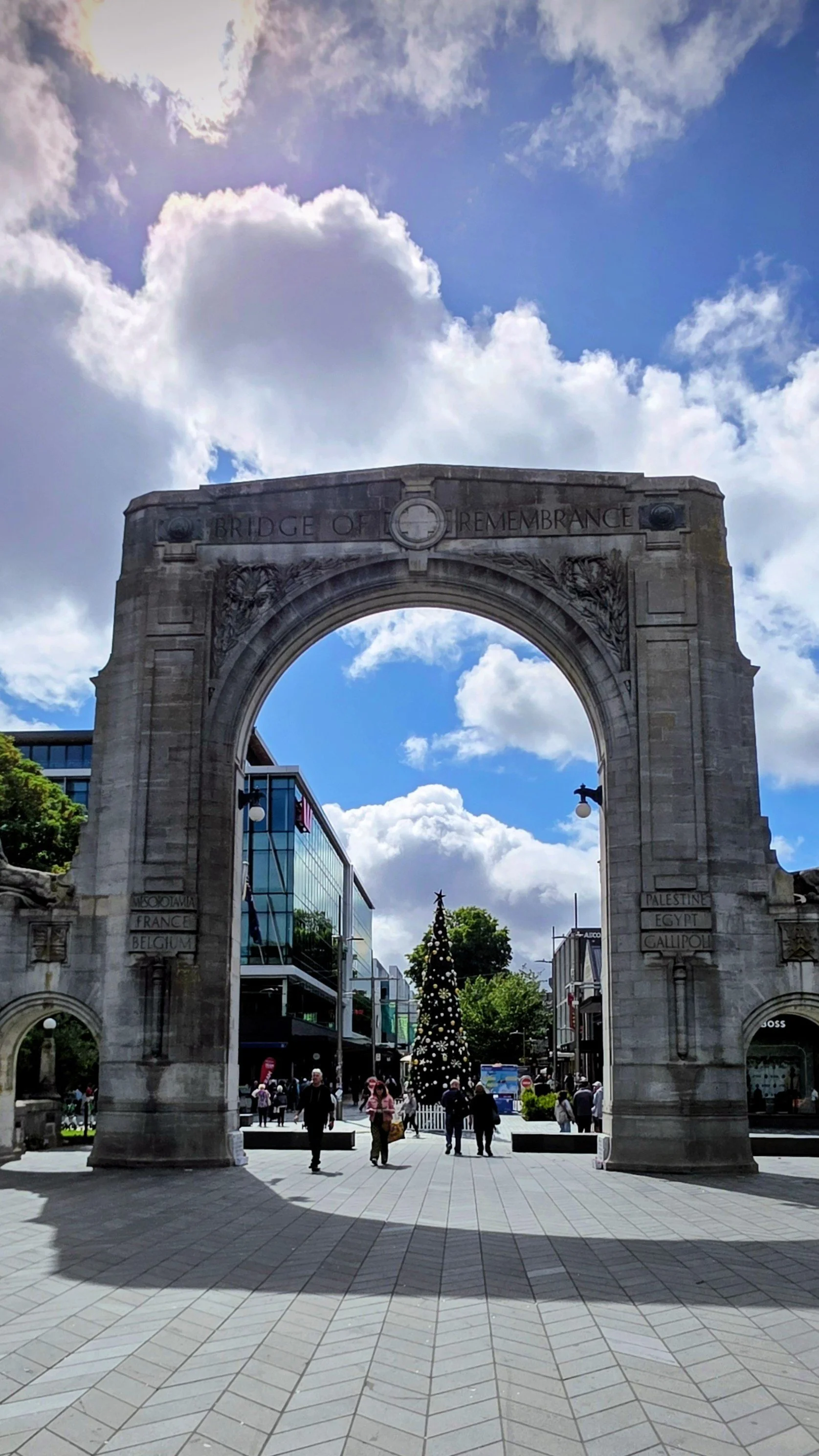 Arch with the words 'Bridge of Remembrance' engraved at the top, leading to a decorated Christmas tree in a busy shopping area with people walking and modern buildings.