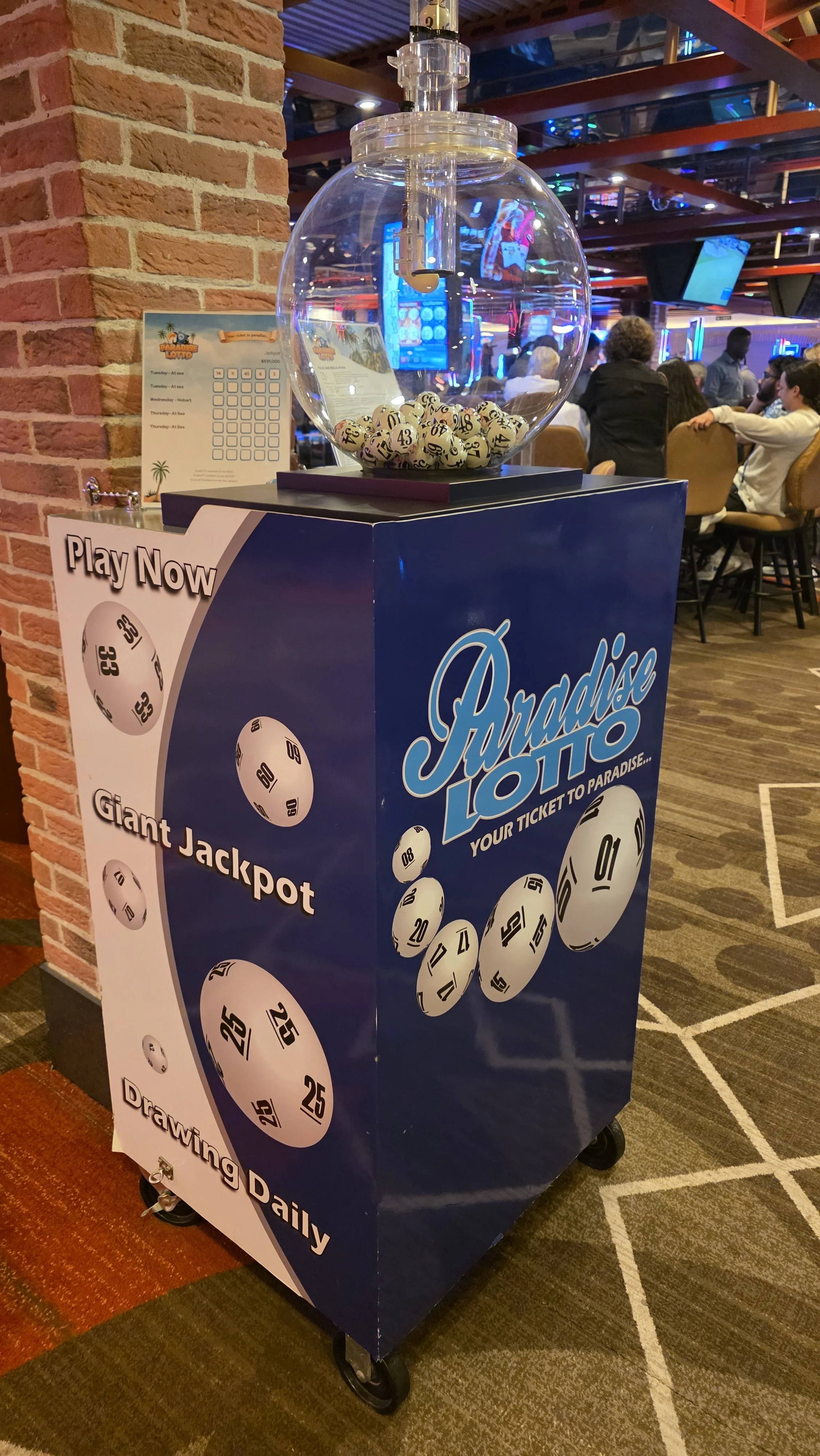 A lottery machine with a transparent globe containing numbered balls, positioned on a blue stand labeled 'Paradise Lotto'. The stand displays images of lotto balls with numbers 25, 15, 20, 08, and 25. The background shows a casino or entertainment ve