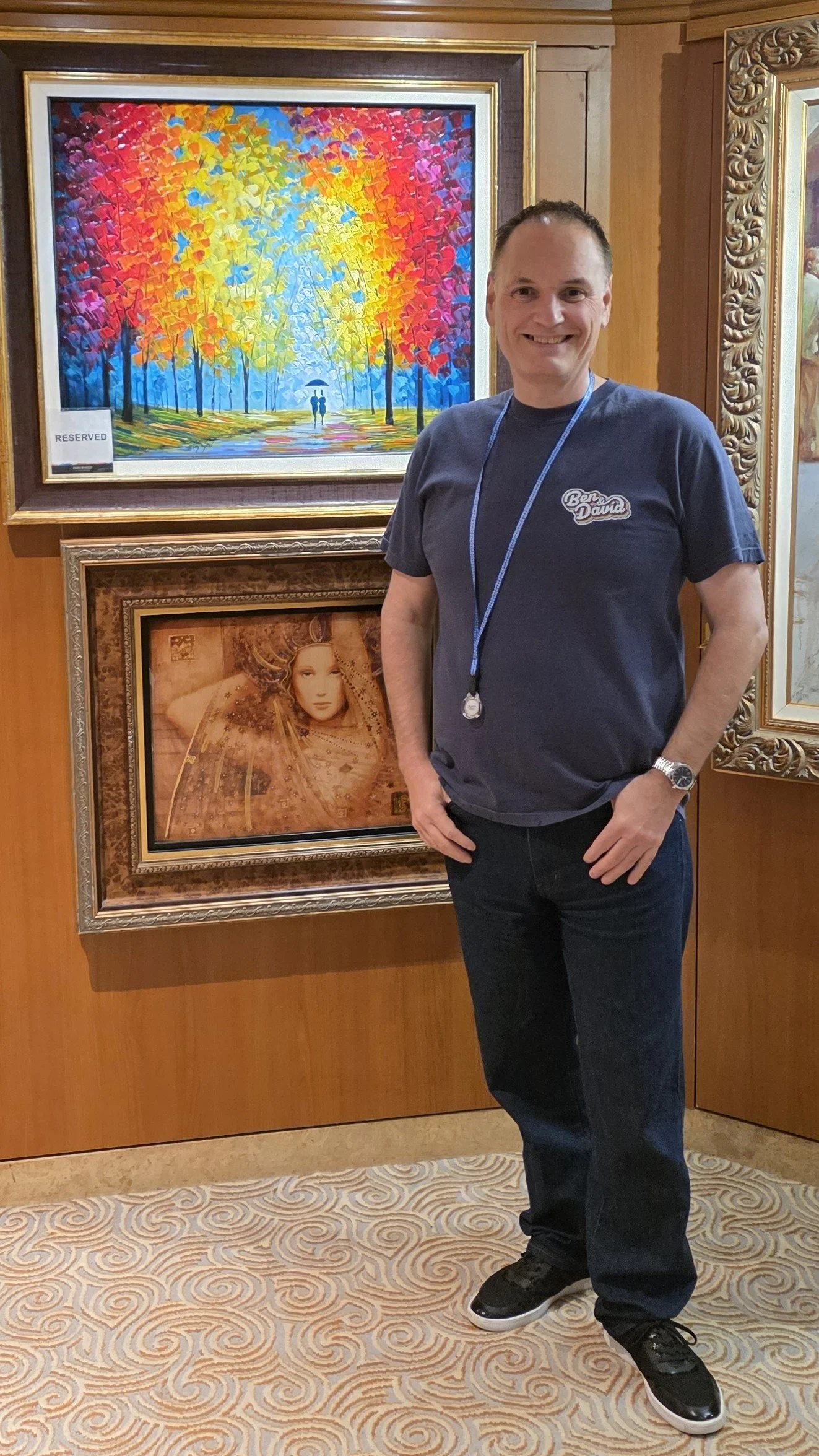A man standing in front of colorful framed paintings on a wall, with a wood-paneled wall behind him and a patterned carpet below.