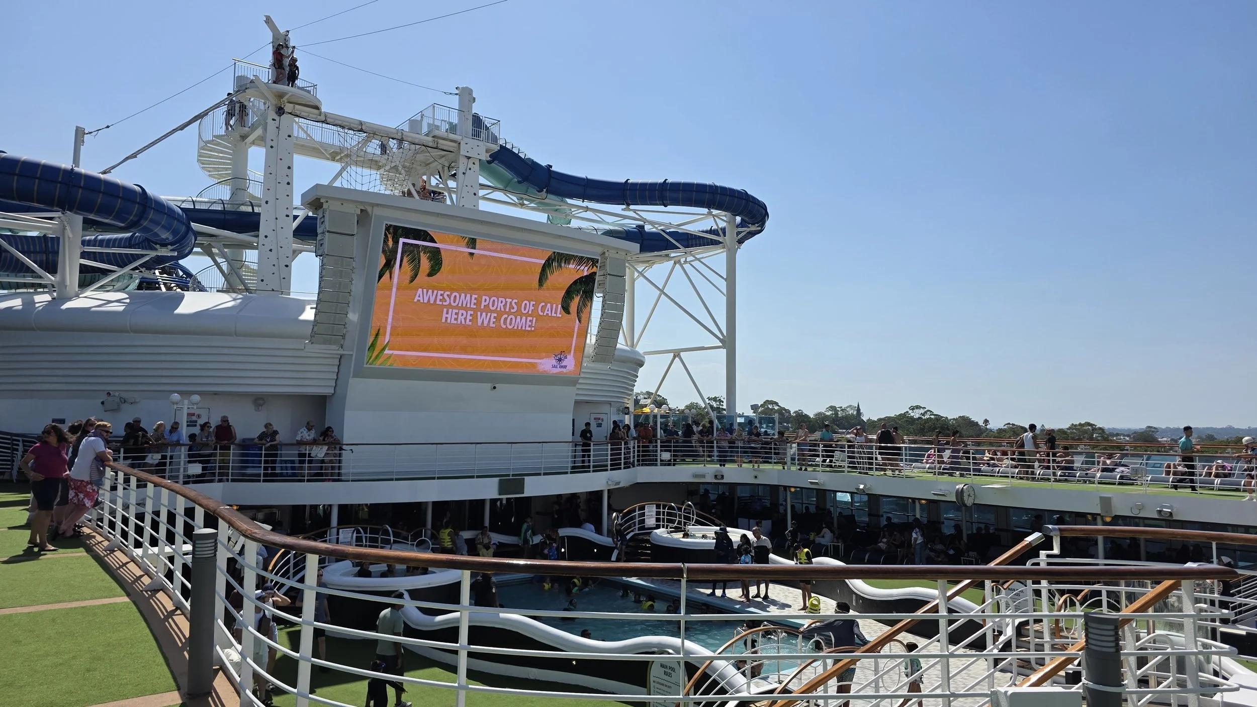Cruise ship deck with pool, waterslides, and large digital screen displaying a tropical scene with palm trees and the text 'Awesome ports of call, here we come!' blue sky and passengers walking and relaxing.