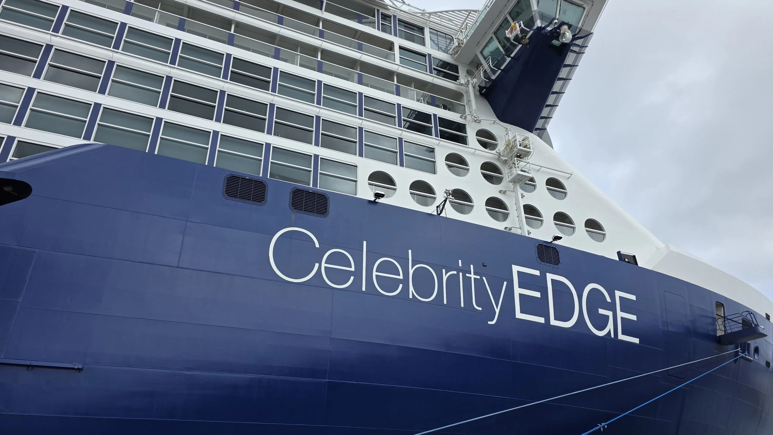Close-up of a cruise ship named 'Celebrity Edge' with multiple windows and circular portholes, docked outdoors under cloudy skies.
