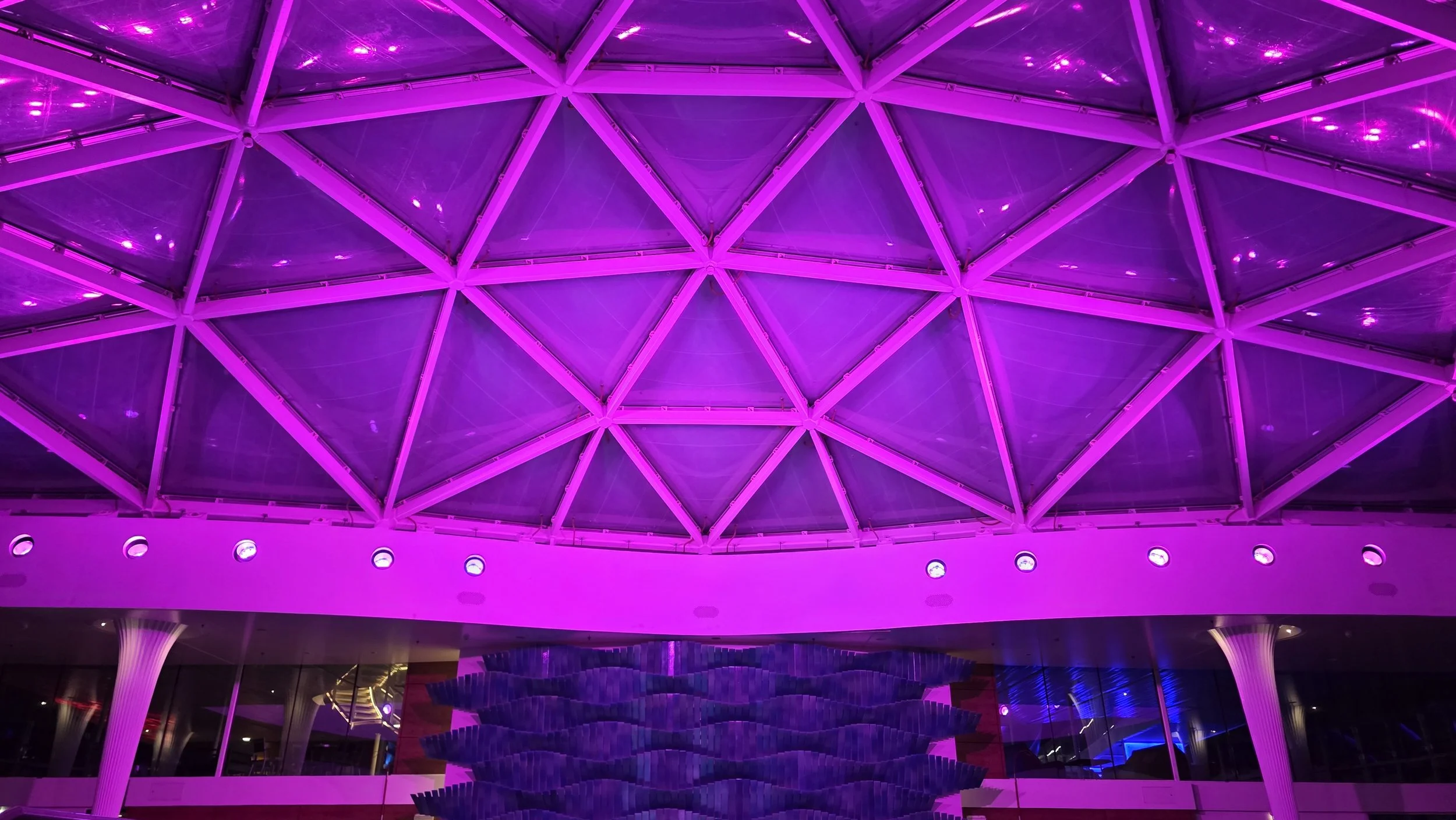 Interior of a modern building illuminated with purple and pink lighting, featuring a geometric, triangular-patterned ceiling and large, white column supports.