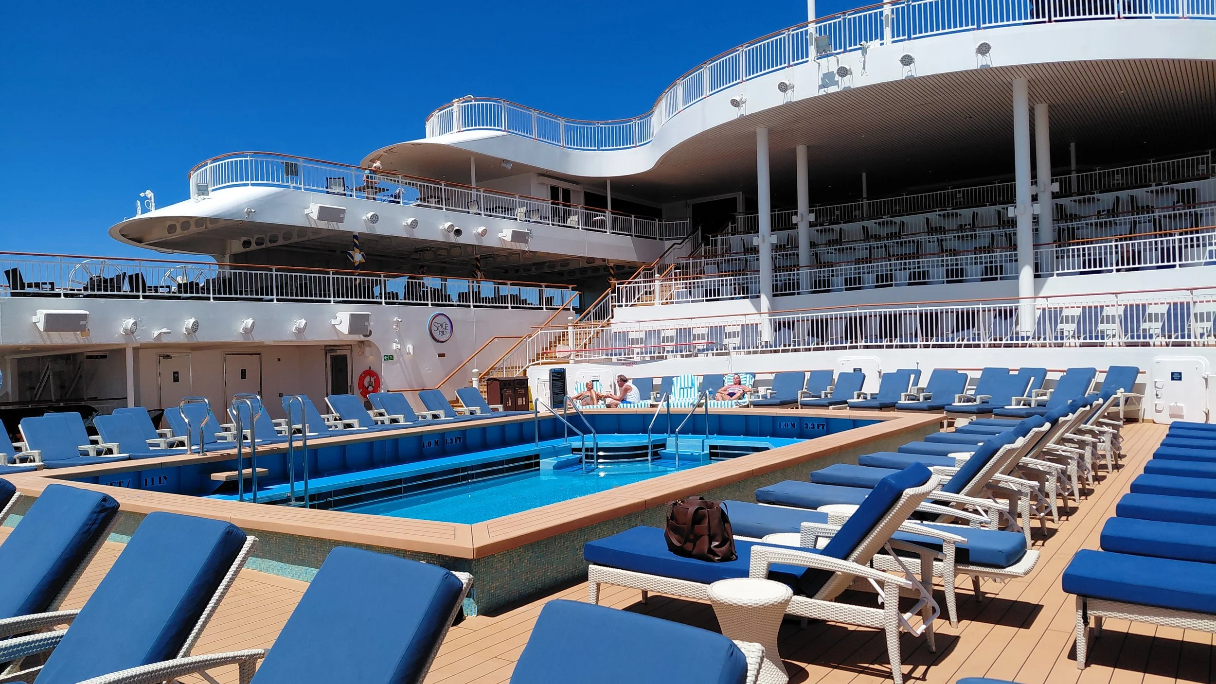 Empty swimming pool on a cruise ship deck with blue lounge chairs, surrounded by a multi-level open-air deck and railings under a clear blue sky.