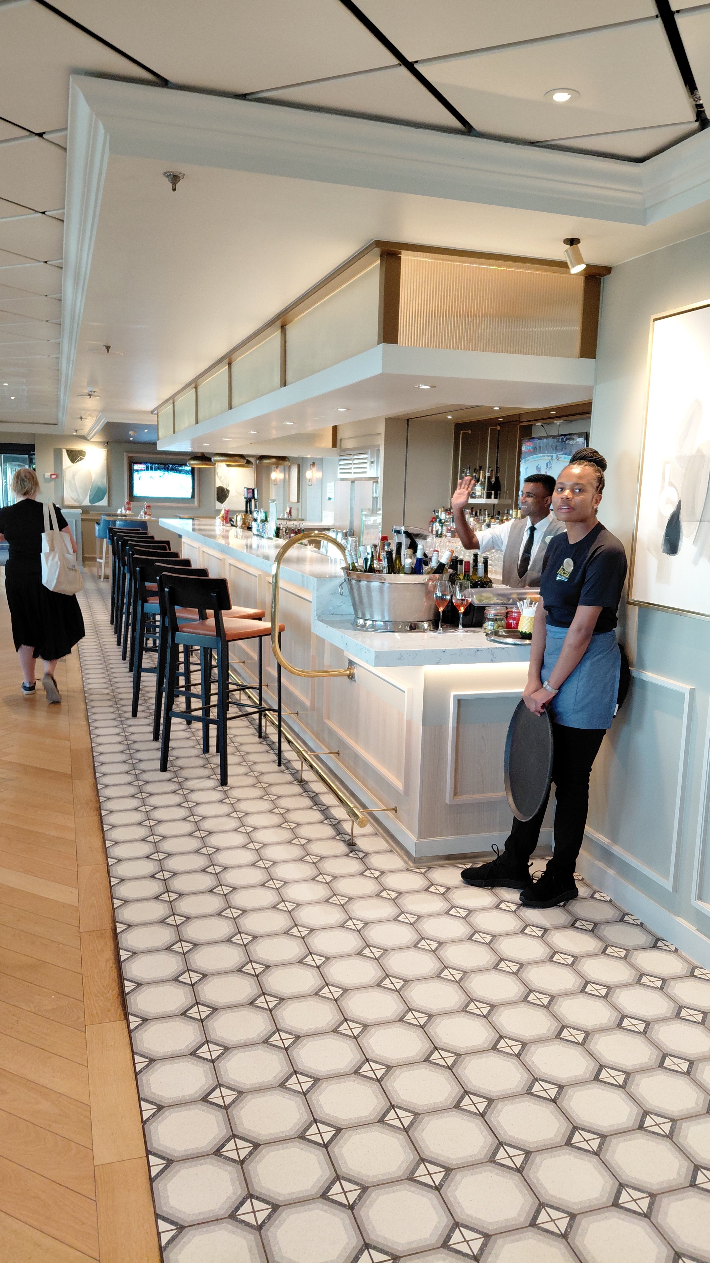 Inside a modern bar with two bartenders, one waving and the other holding a tray. The bar has a white marble counter with barstools, and a woman is walking away in the background. The floor has patterned tiles and the ceiling has a mix of flat and dr