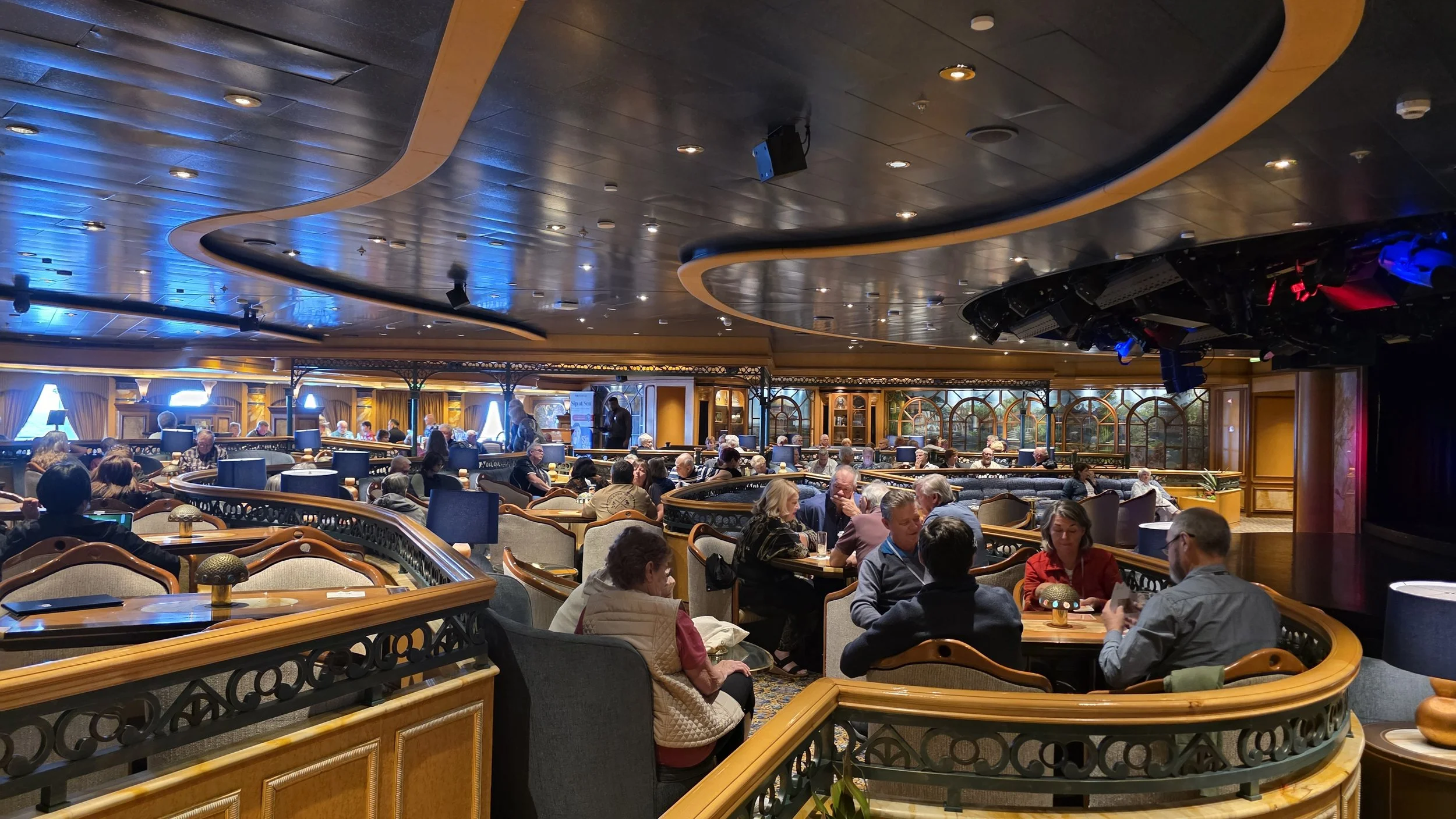 Interior of a lively restaurant or lounge with patrons sitting at tables, ornate woodwork, large arched windows, and a ceiling with recessed lighting.