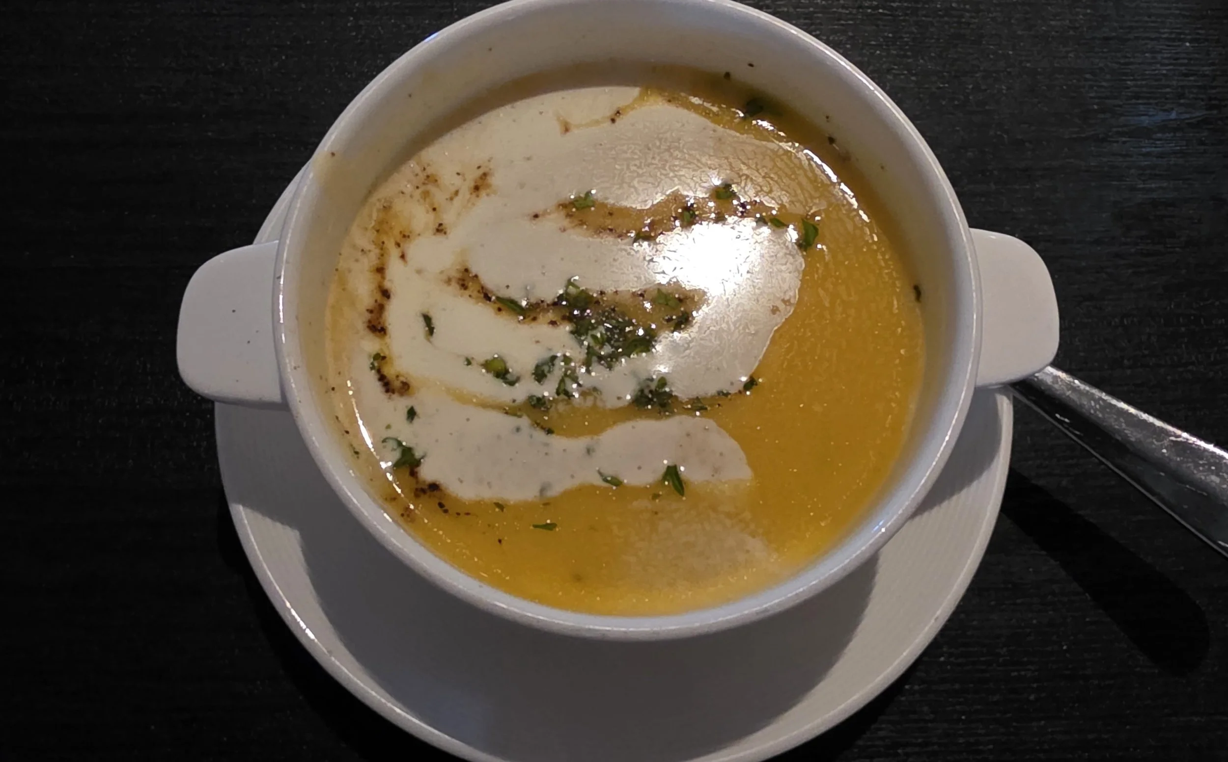 A bowl of creamy soup garnished with herbs, served on a white plate with a spoon on a dark wooden table.