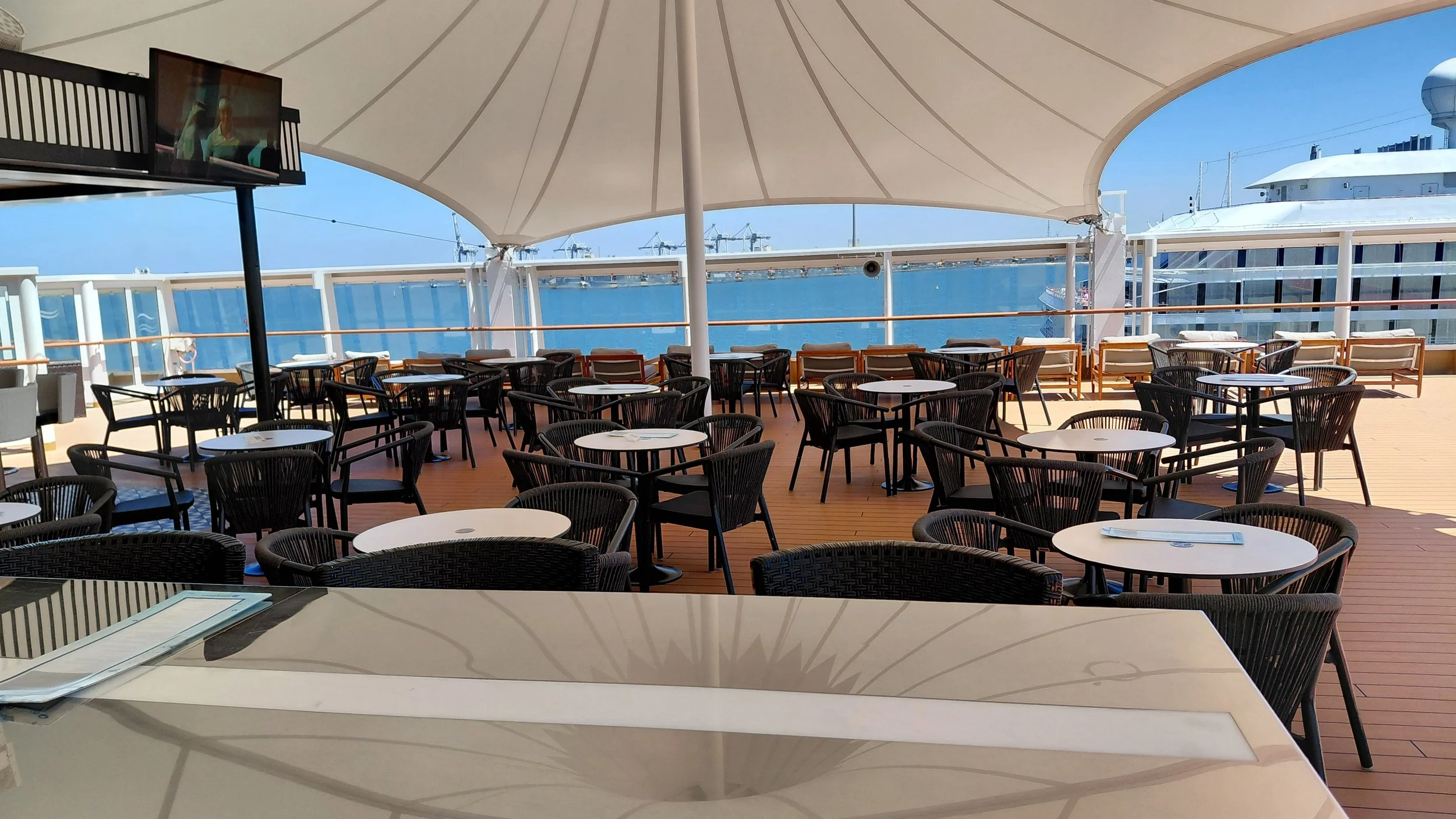 Outdoor seating area on a cruise ship deck with tables and chairs, covered by a large sail, overlooking the ocean with a yacht and cranes in the background.