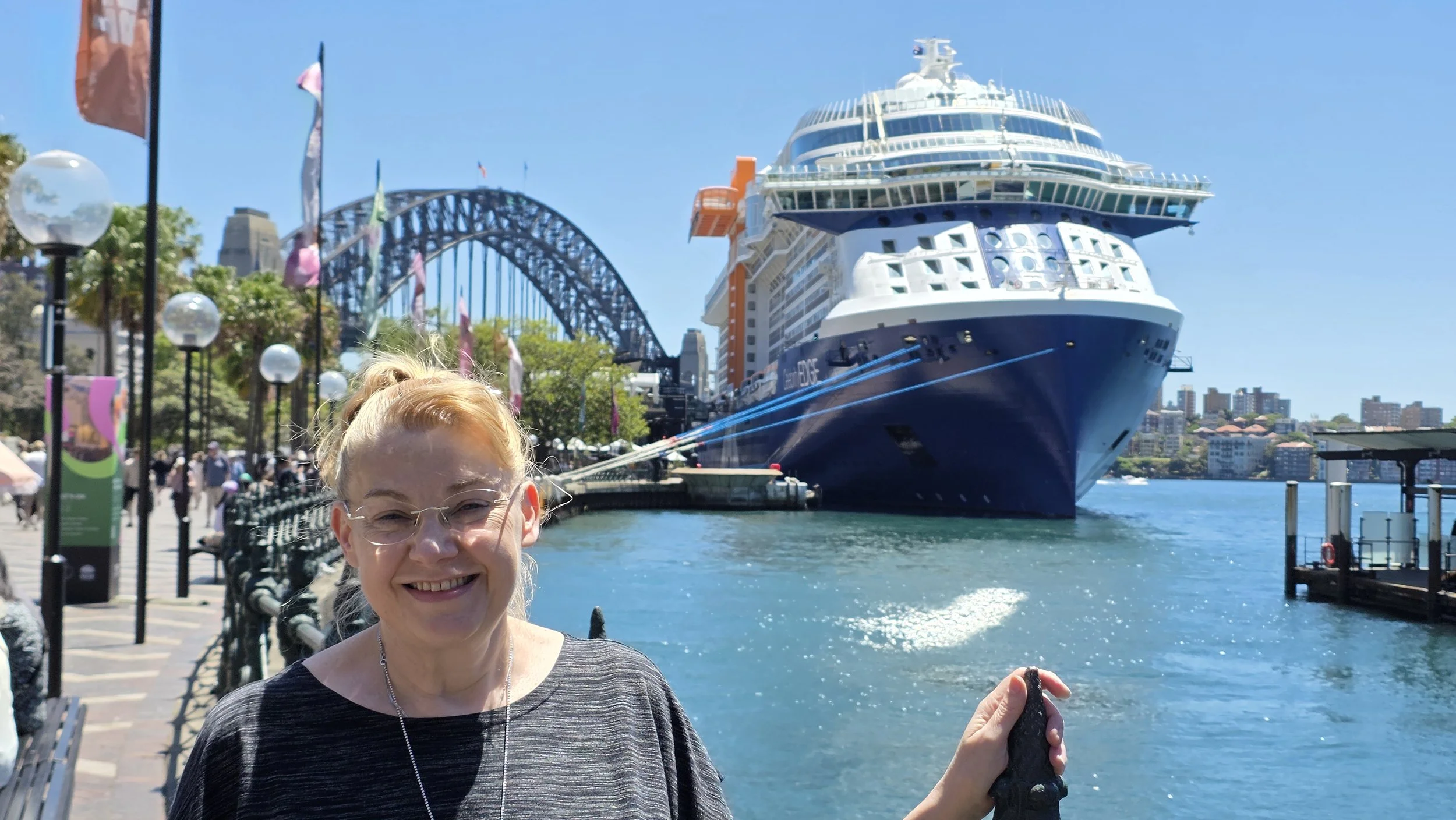A smiling woman with glasses and blonde hair standing near the water in Sydney, Australia, with the Sydney Harbour Bridge and a large cruise ship in the background on a sunny day.