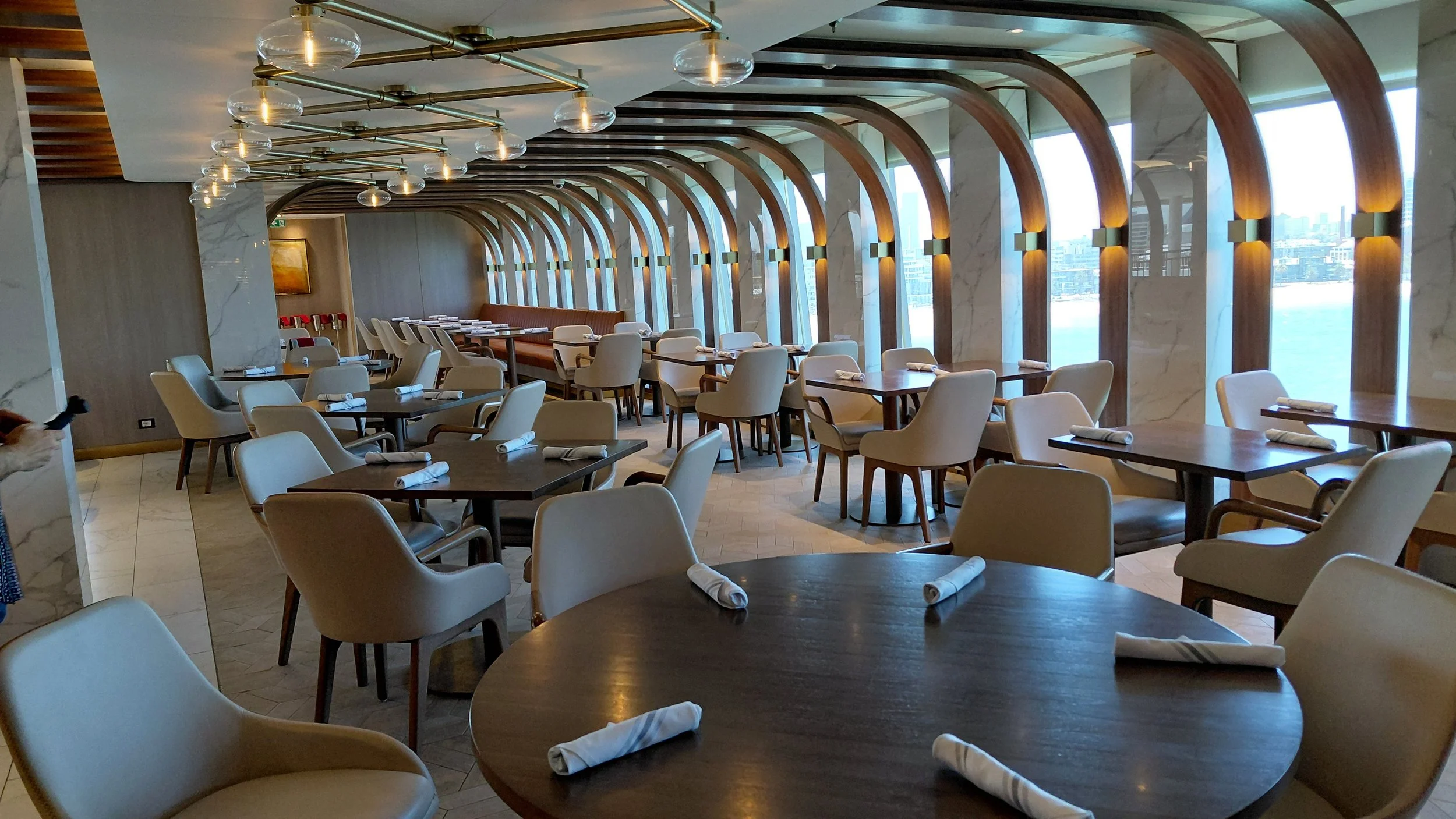 Elegant restaurant interior with large arched windows offering a view of the city and water, modern lighting fixtures, arranged tables with napkins, and comfortable chairs.