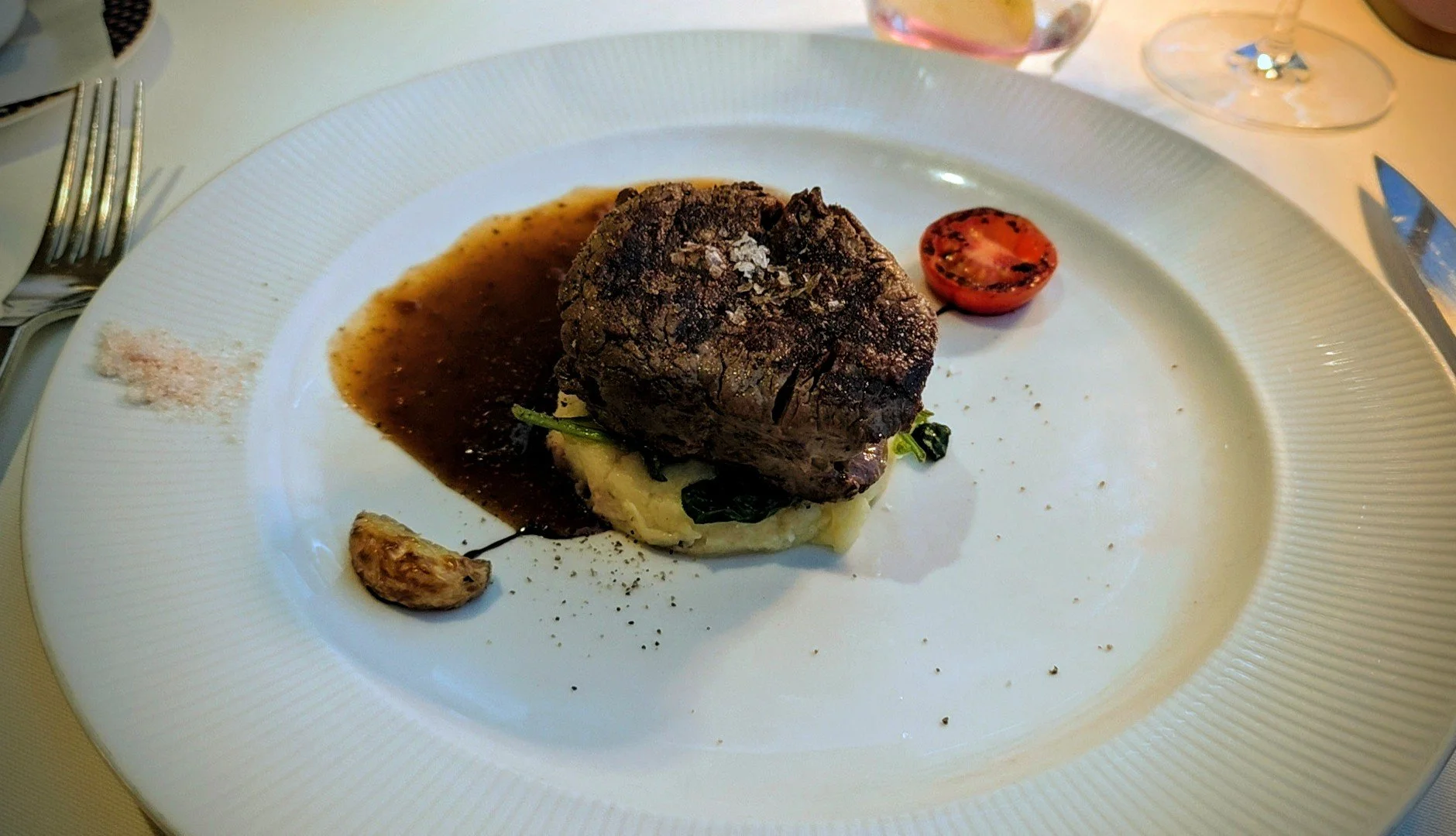 A plated meal consisting of a beef steak topped with coarse salt, served with mashed potatoes, green vegetables, a roasted garlic clove, and a side of dark gravy, with a halved cherry tomato on the side.