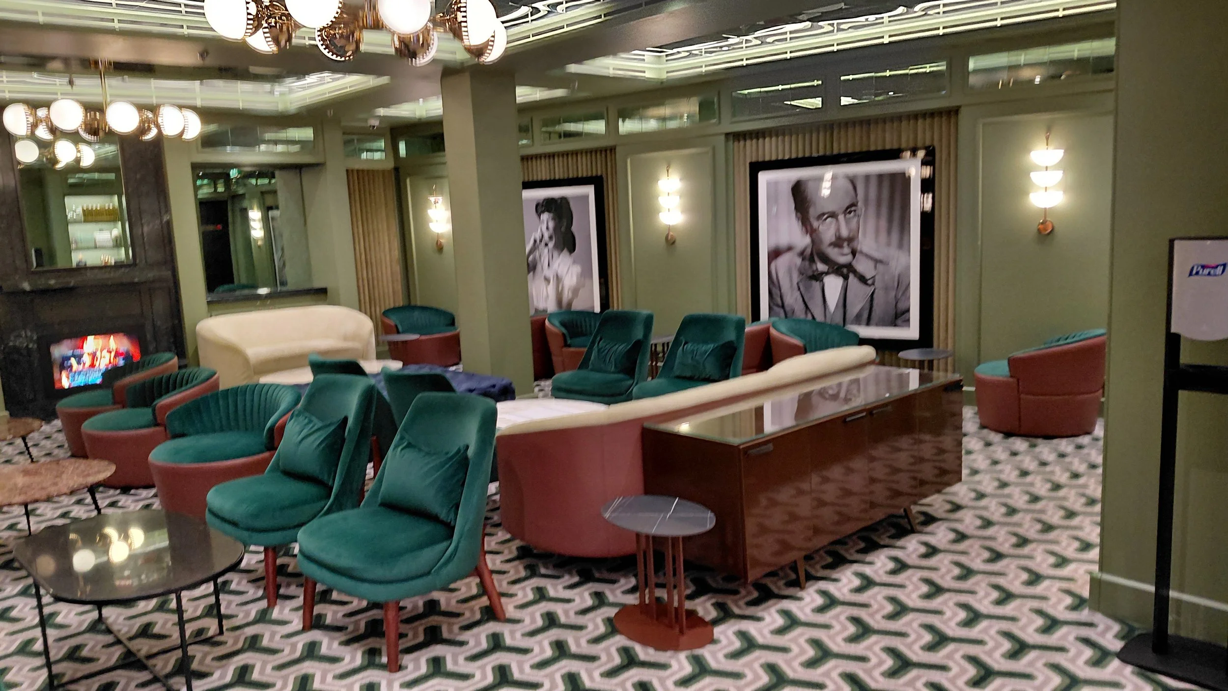 Interior of a vintage-style lounge with green and pink chairs, a white sofa, and black-and-white framed portraits on the walls.