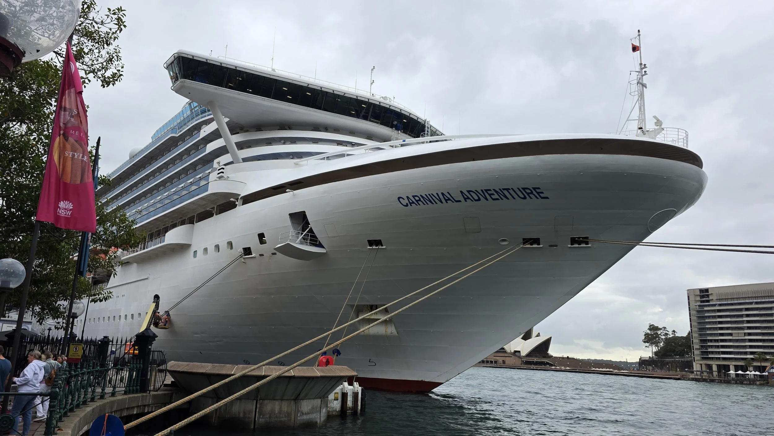 A large white cruise ship named Carnival Adventure docked at a harbor with people walking on the pier and buildings in the background.