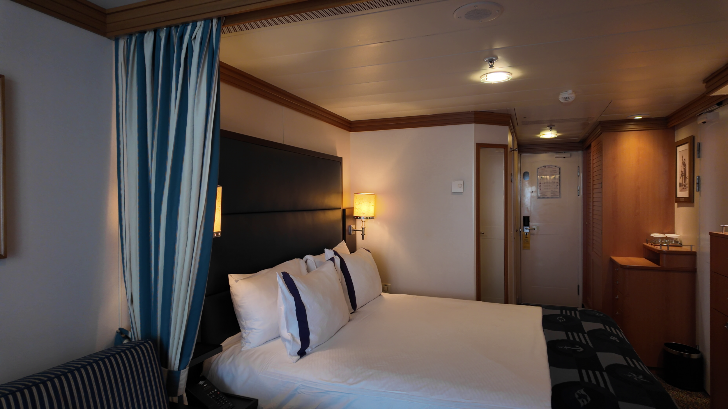 Interior of a hotel room with a bed, pillows, nightstand, lamps, curtains, and wooden furniture near the entrance.