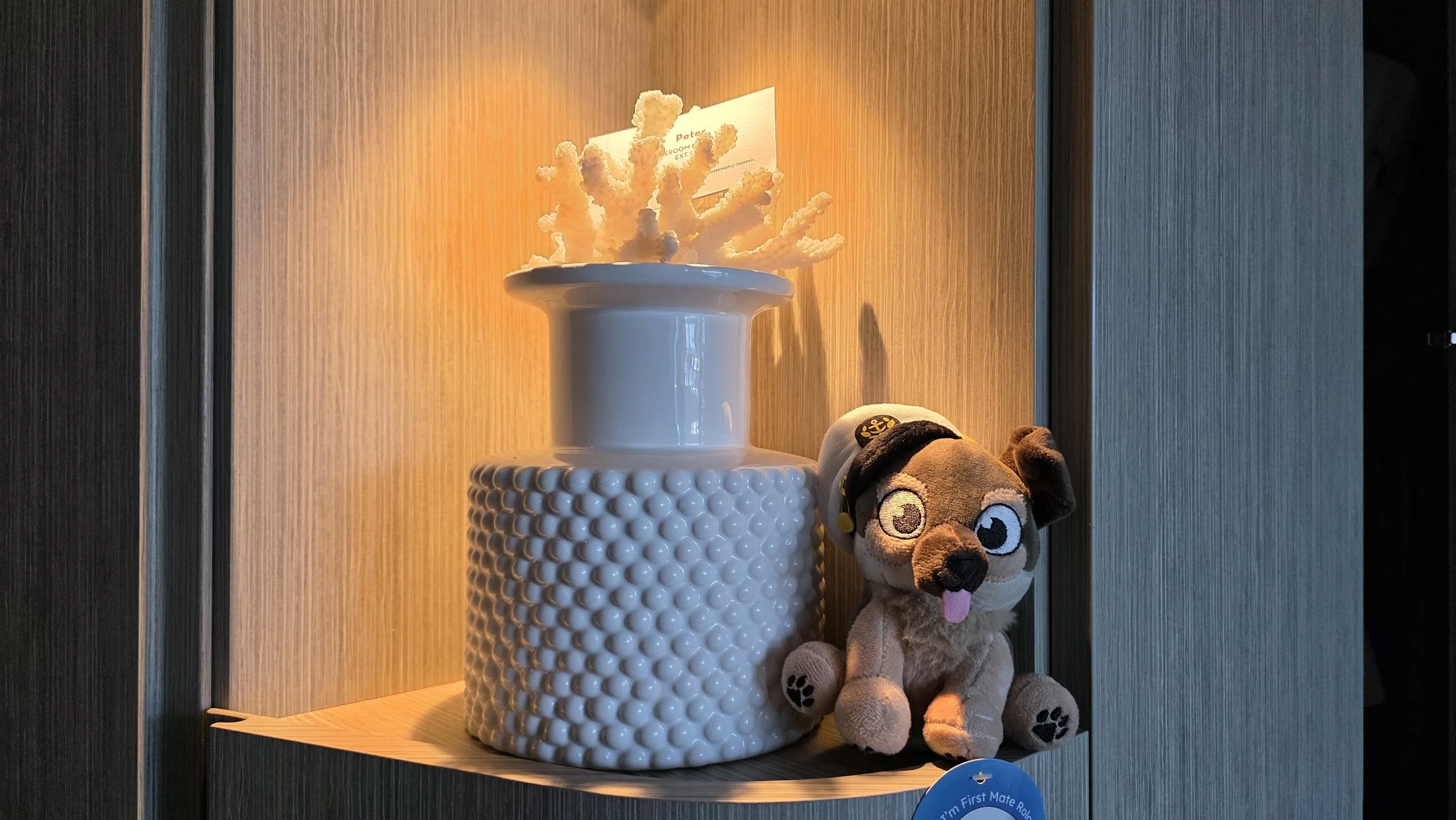 Decorative shelf with a white textured vase emitting a warm glow and a plush dog toy wearing a hat, placed in a wooden niche.