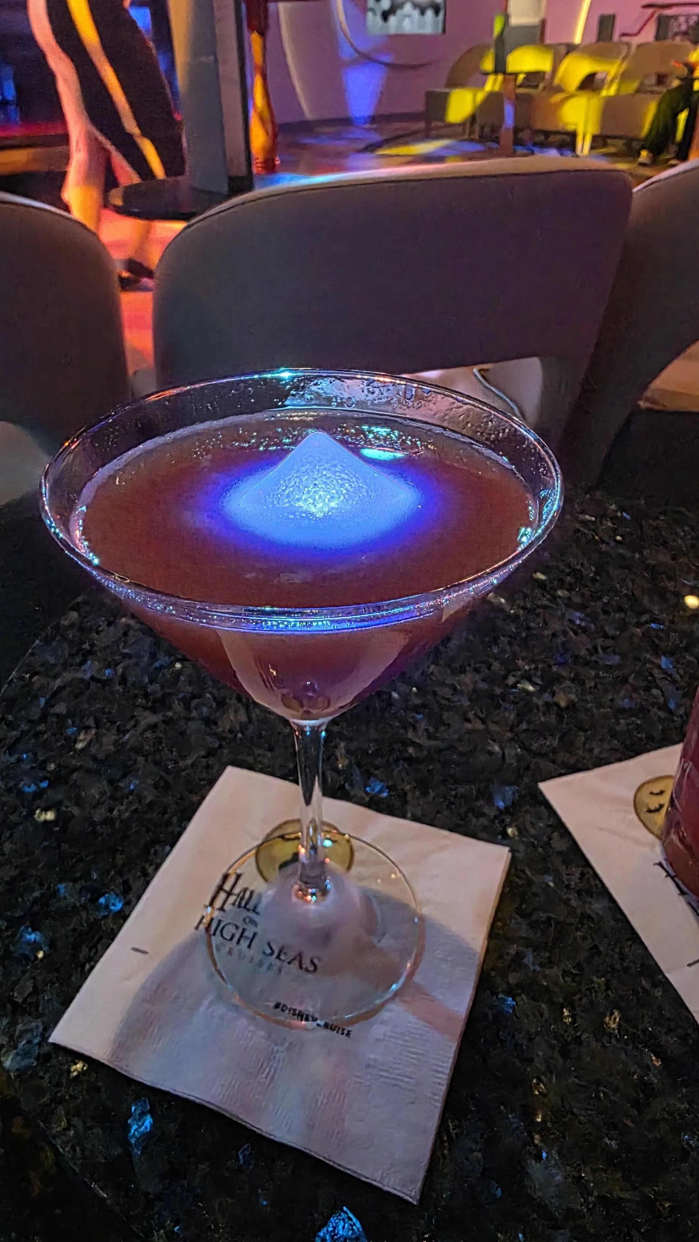 A cocktail glass with a pink drink and a large ice cube, placed on a napkin that has the text 'HILL HIGH SEAS' on it, in a bar or nightclub setting with colorful lighting.