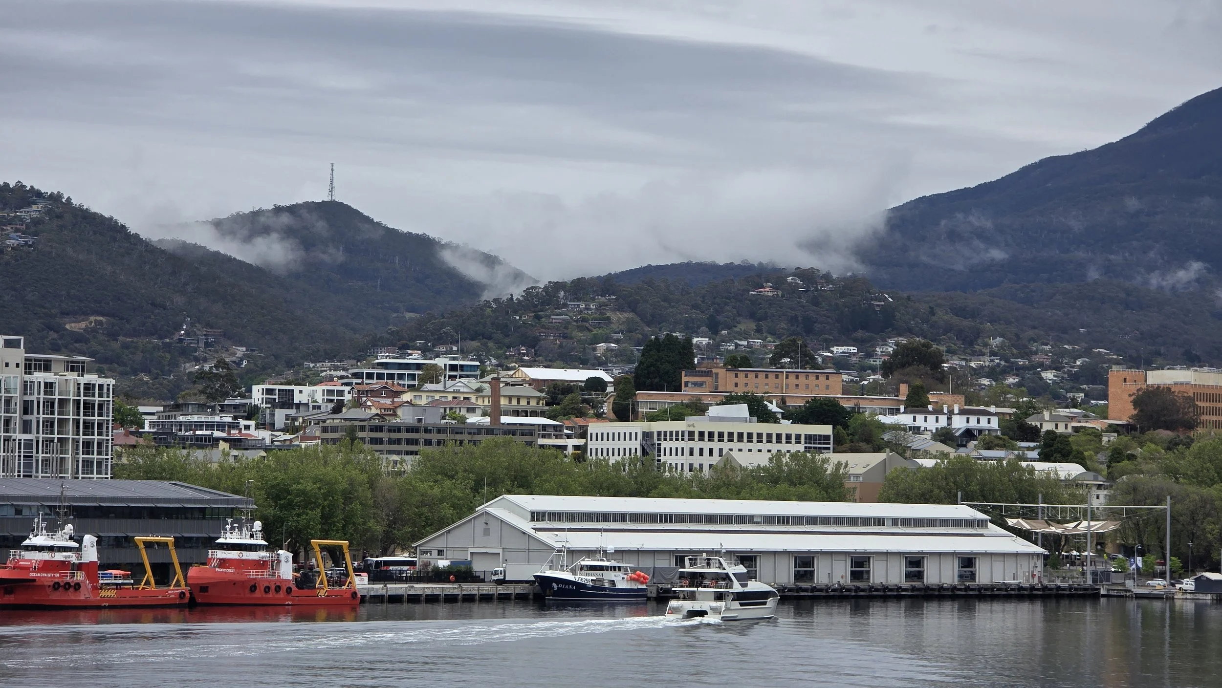 View of a harbor with boats docked, a mountain range in the background, and a cloudy sky overhead.