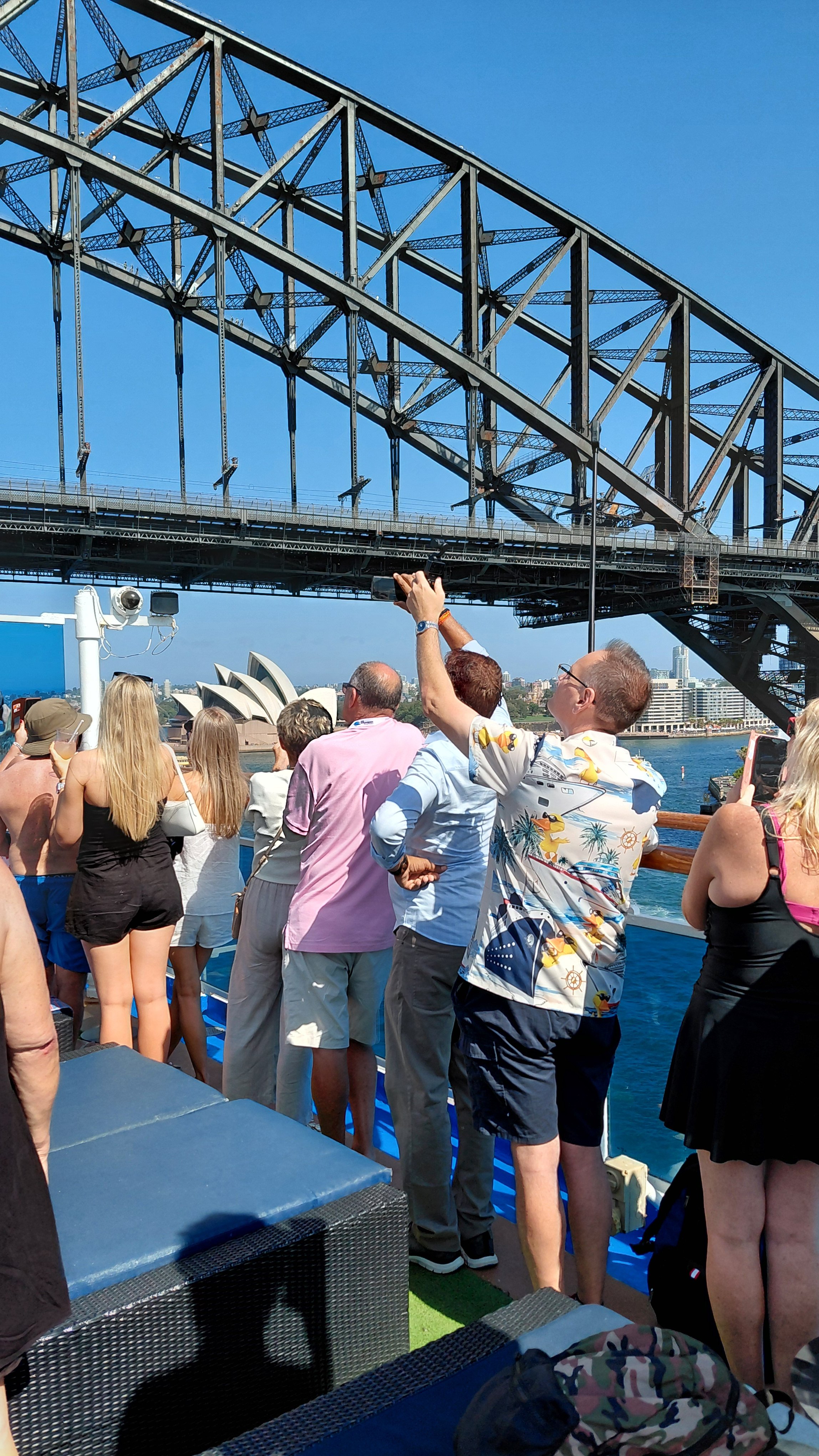 People on a boat taking photos and enjoying a sunny day with the Sydney Harbour Bridge and Opera House in the background.