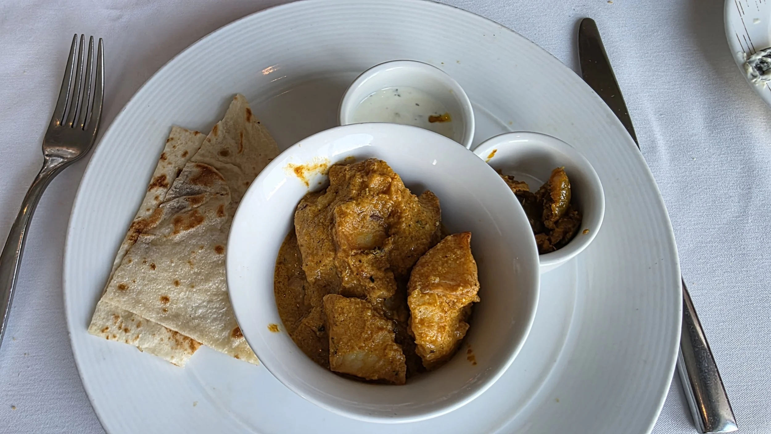 A white plate with naan bread, a bowl of chicken curry, two small cups of condiments or sauces, and cutlery on a white tablecloth.