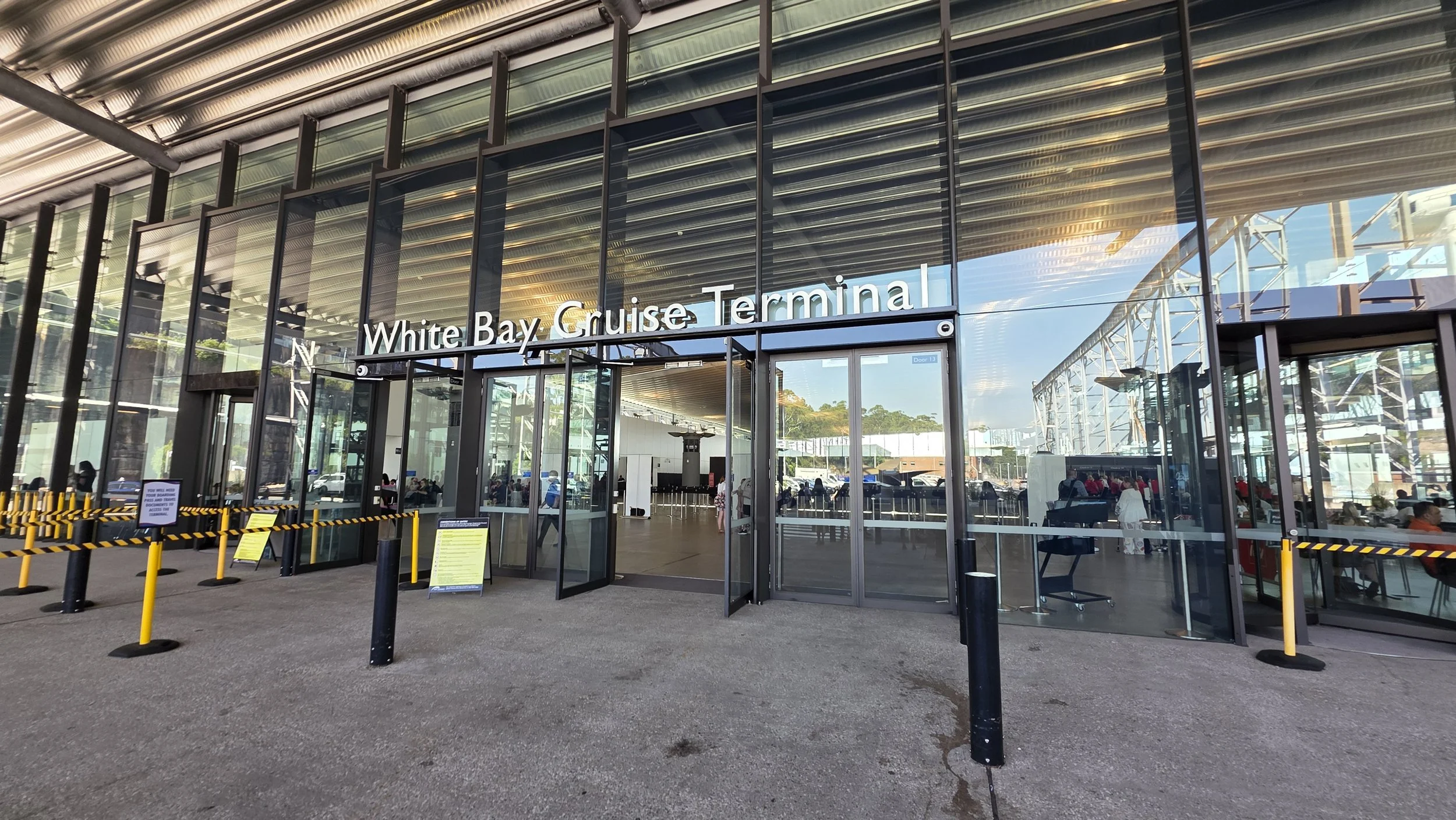 Front view of the White Bay Cruise Terminal with glass doors and windows, people sitting and walking inside, and traffic outside, during daytime.