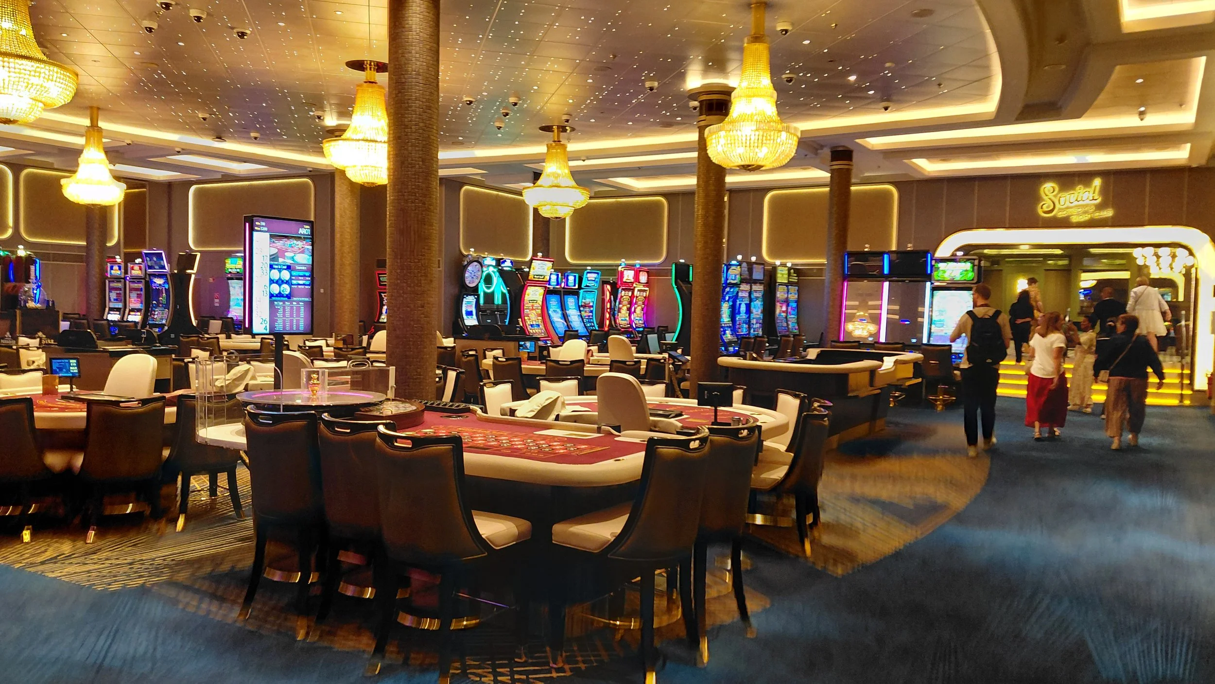 Casino interior with slot machines, electronic gaming tables, chandeliers, and people walking near the entrance with a 'Social' sign. The room features a luxurious decor with gold accents and warm lighting.